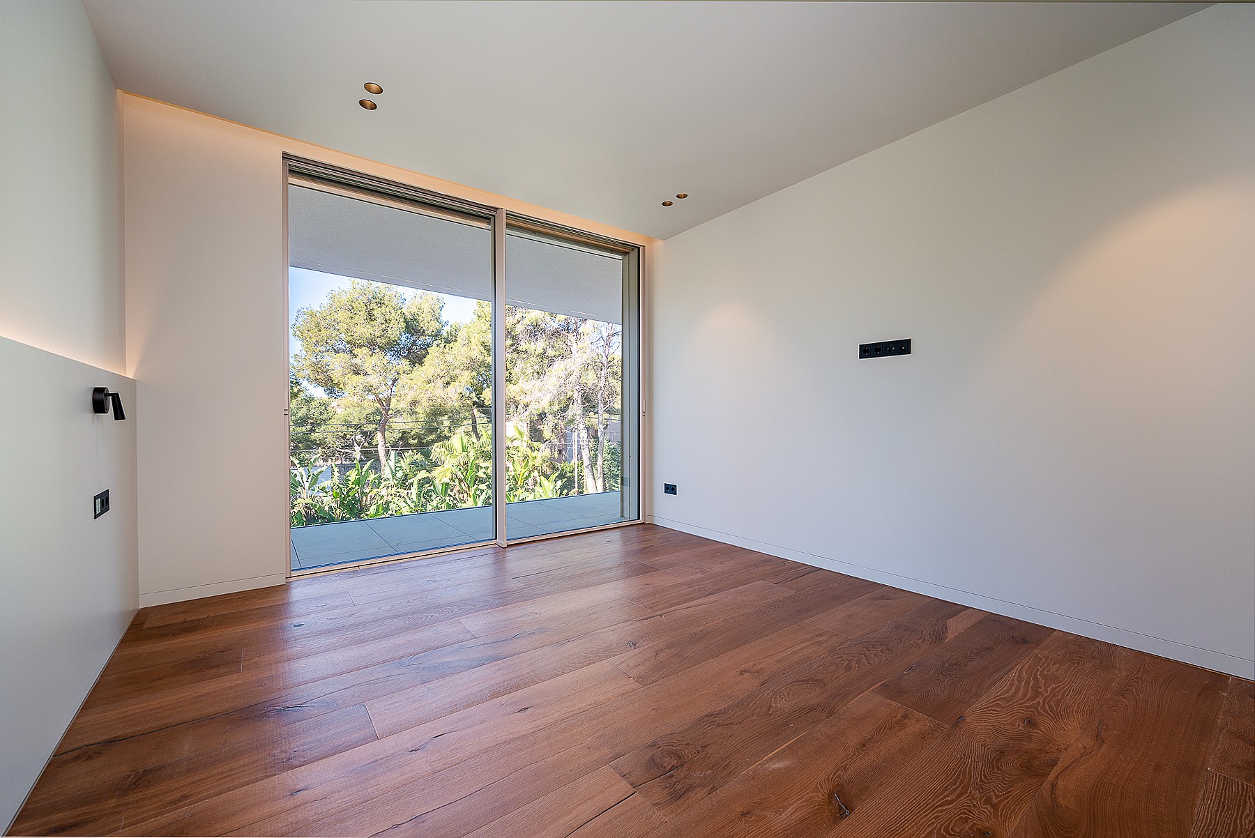 Empty bedroom with wide-plank oak flooring, white walls, recessed ceiling lights, and large sliding glass door to terrace.