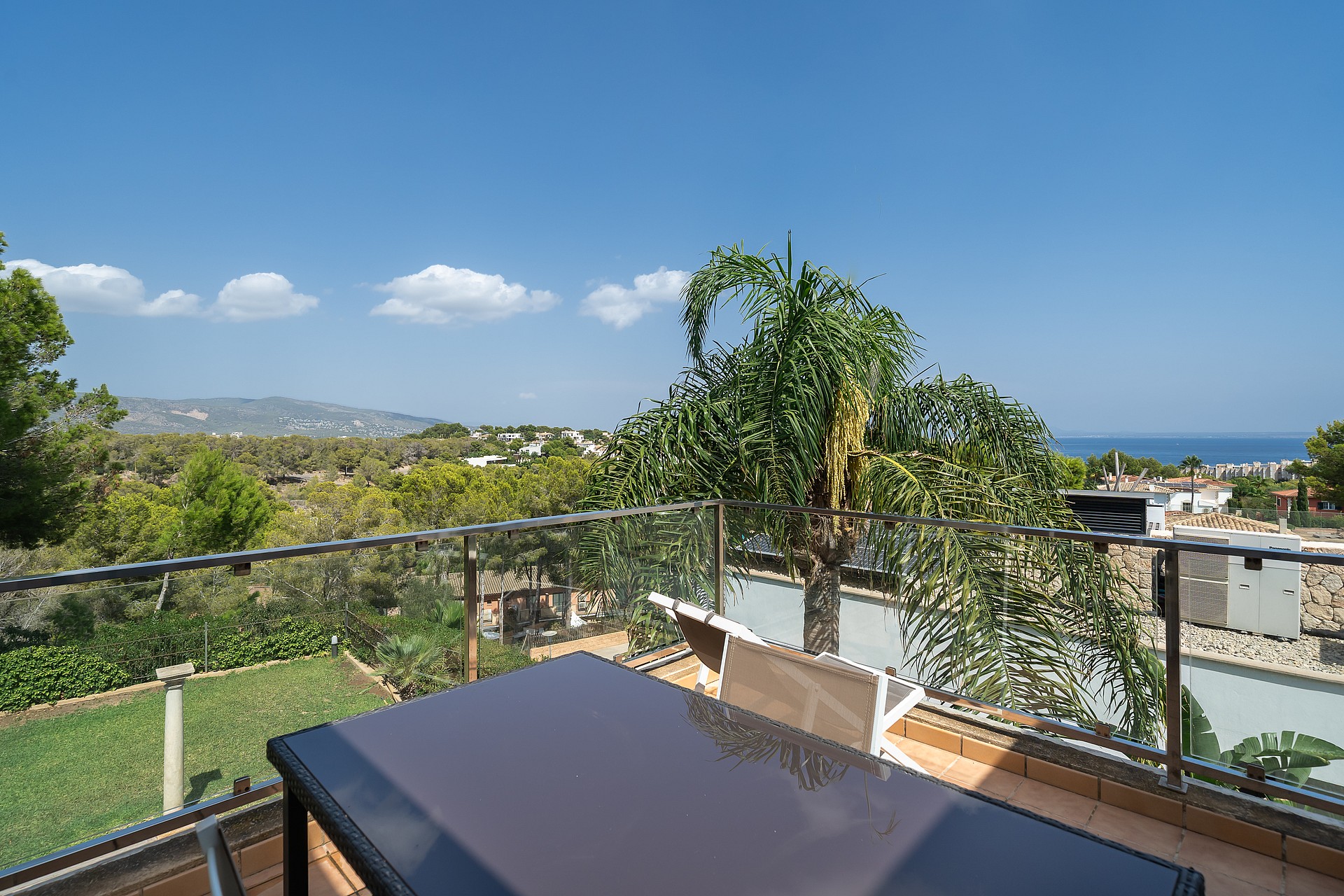 An elevated terrace with a dark outdoor table, glass railing, and distant views of the sea and green landscape.