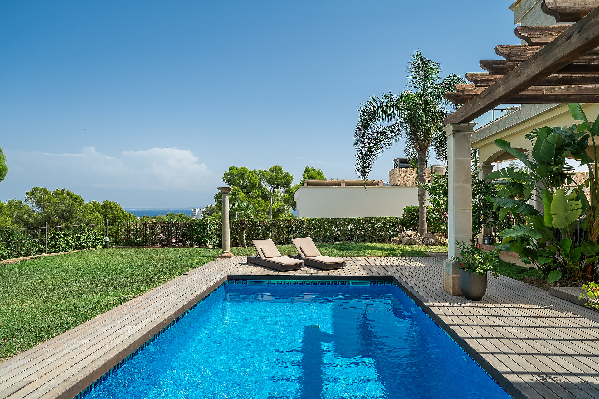 Rectangular swimming pool with blue water next to a timber deck, two sun loungers, and a shaded pergola in a sunny garden.