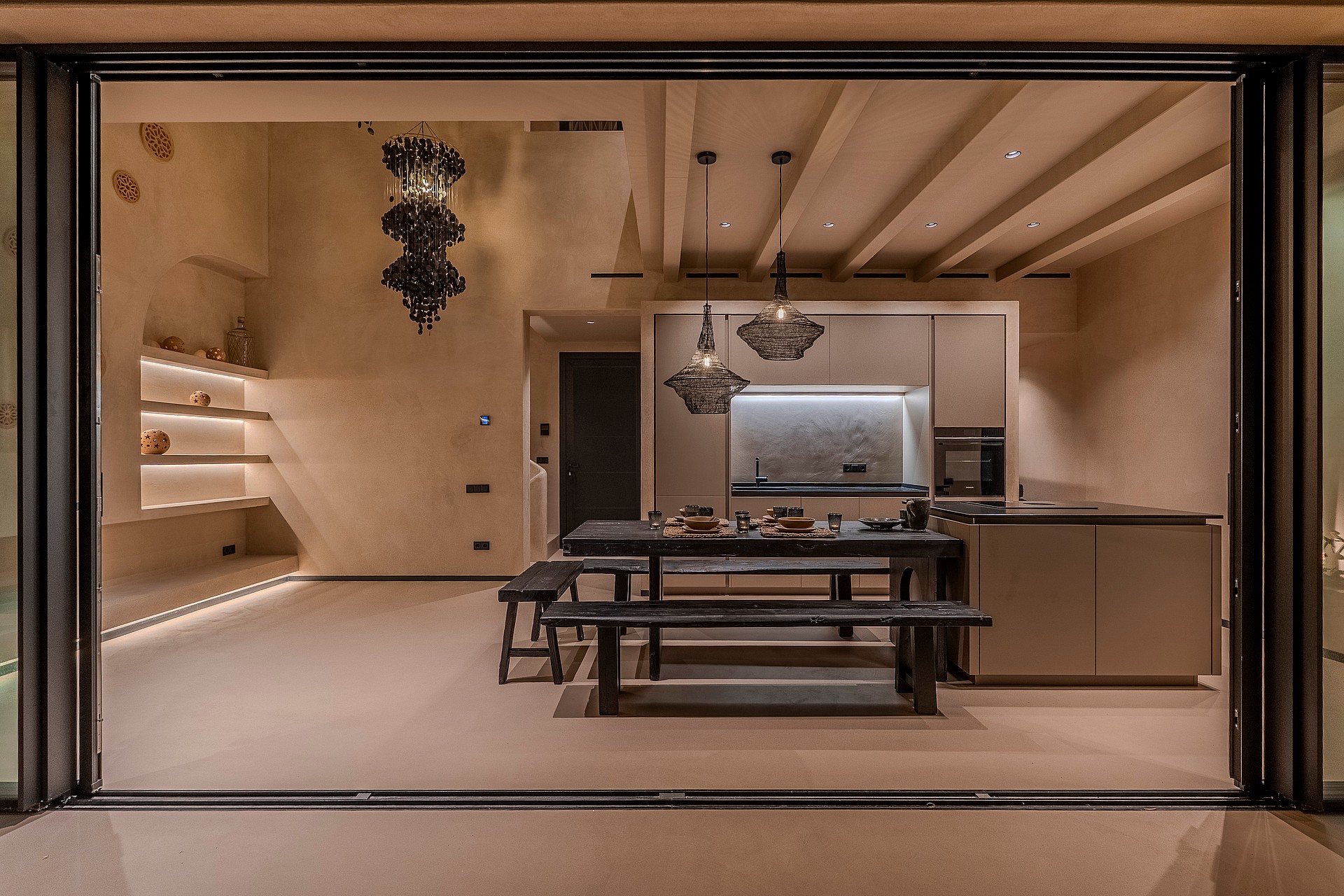 Interior view from a terrace looking into a minimalist kitchen with a wooden dining table, bench seating, and soft recessed lighting.