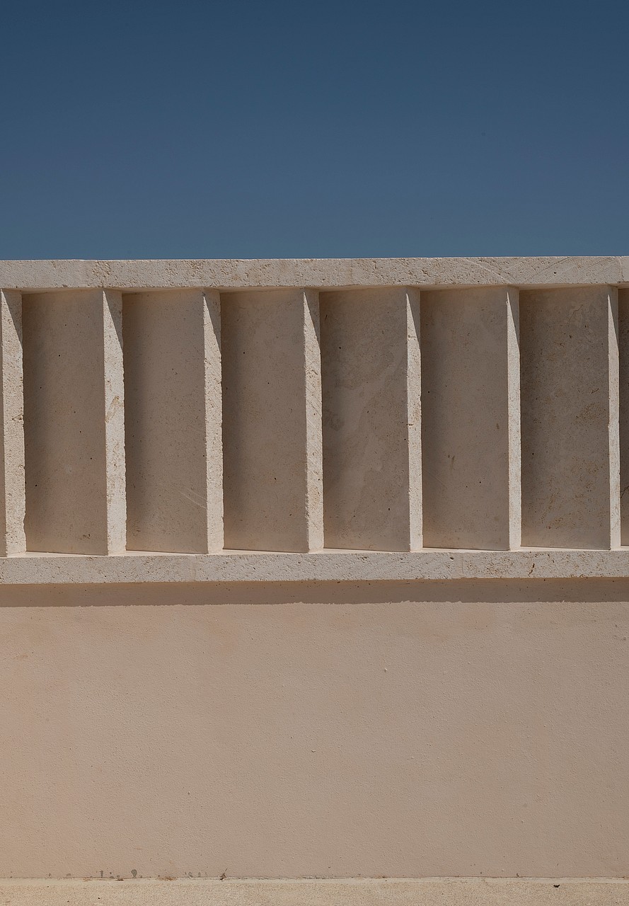 Close-up of a beige sandstone wall with rhythmic vertical recesses creating deep shadows under bright daylight.