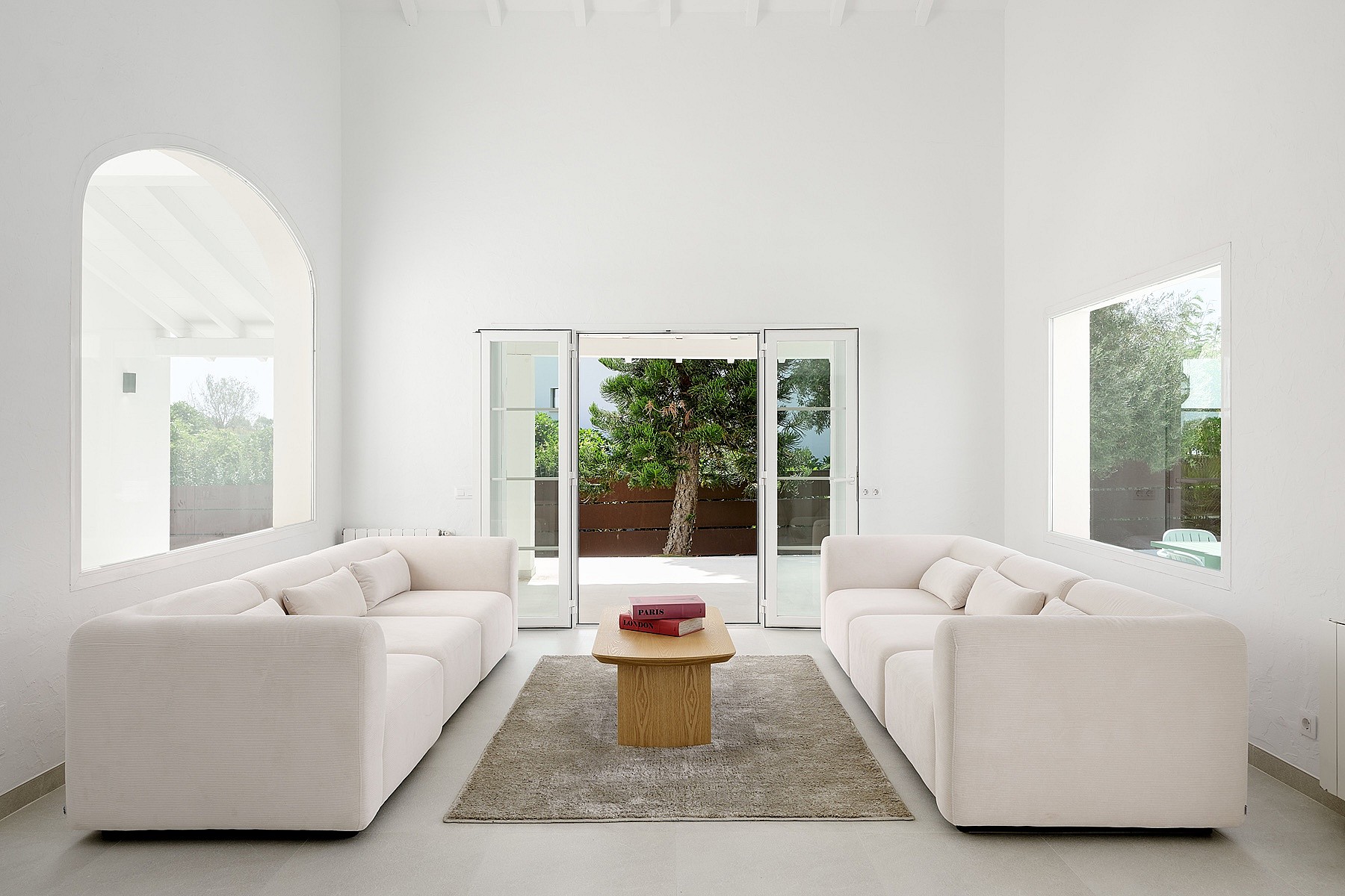 Salón blanco de doble altura con ventana arqueada, dos sofás color crema y puertas acristaladas hacia el jardín