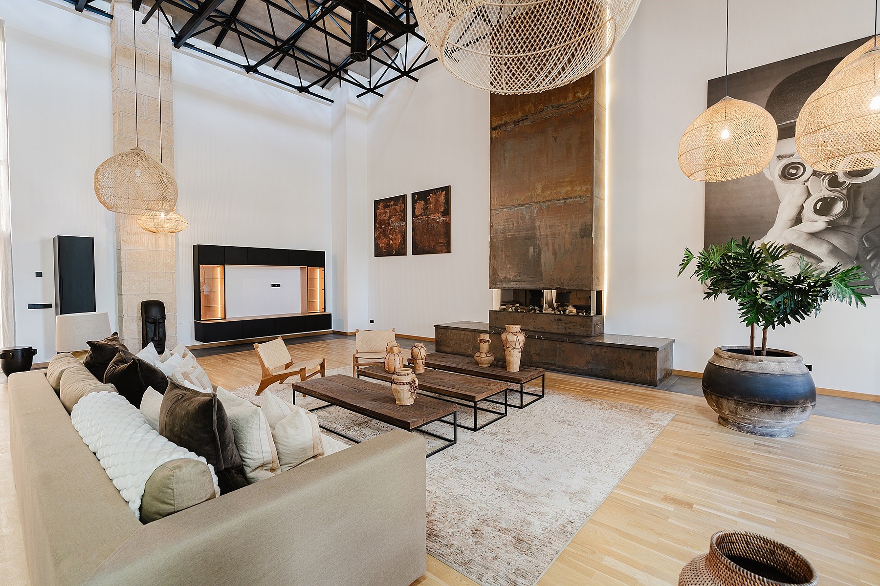 A spacious living area featuring a double-height ceiling with exposed black metal trusses, a tall rusted-metal fireplace surround, and woven pendant lighting.