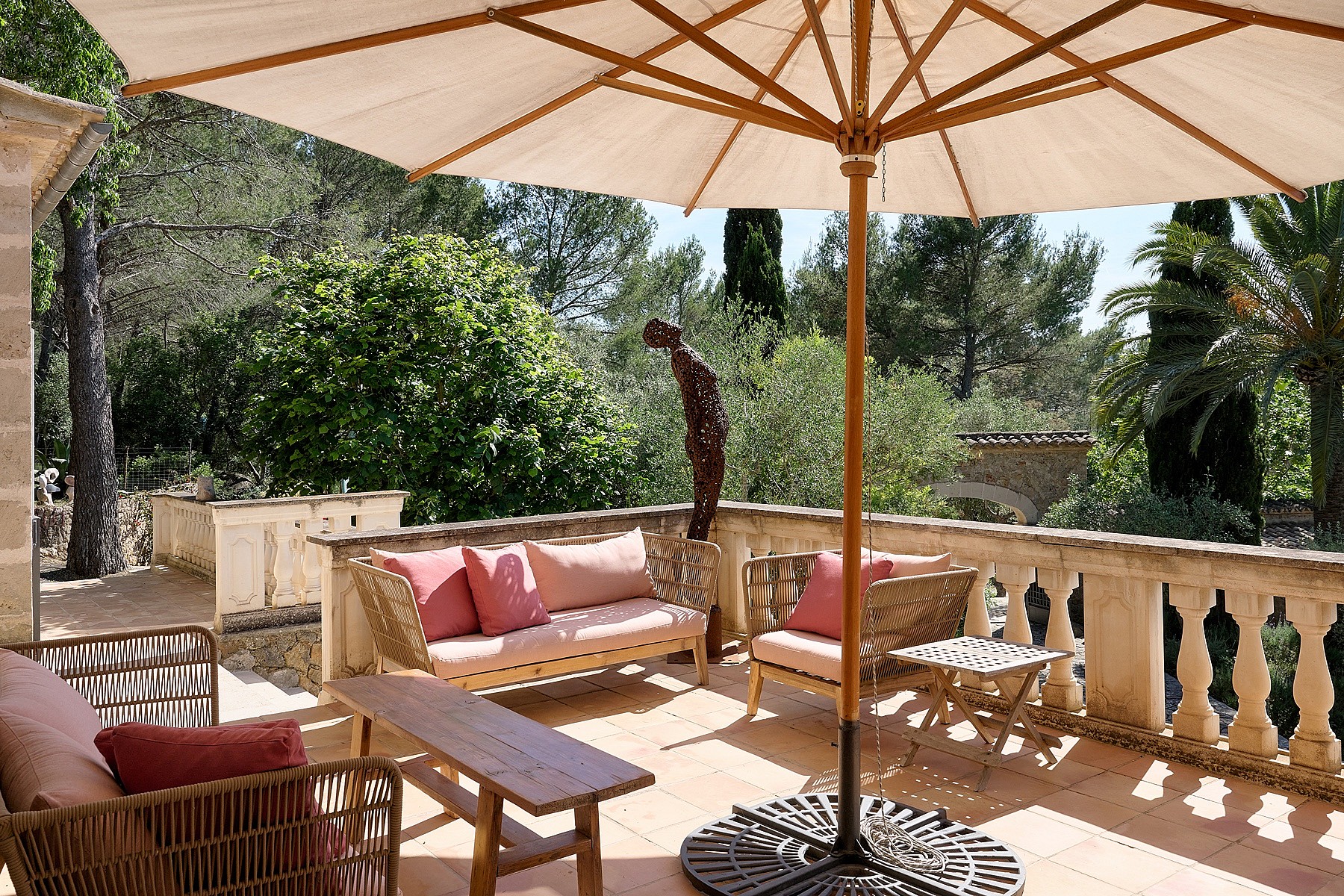A stone terrace with a balustrade, wicker lounge seating with pink cushions, and a large sun umbrella overlooking trees.