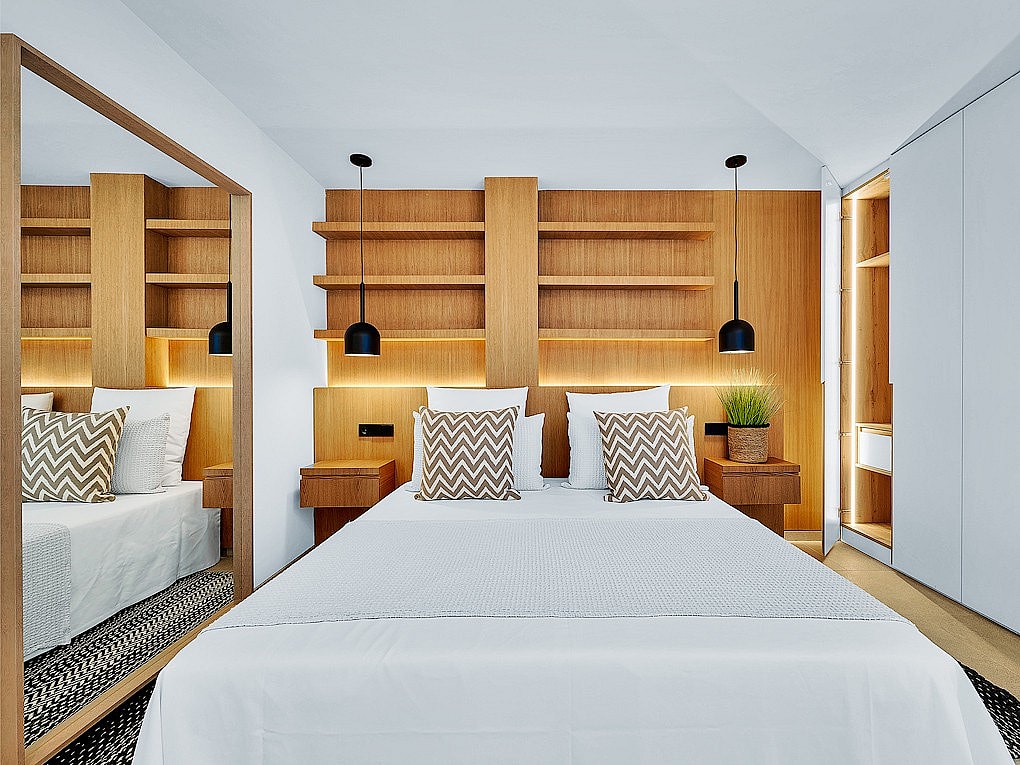A modern bedroom featuring a bed with patterned throw pillows, oak-finished shelving, and integrated lighting.