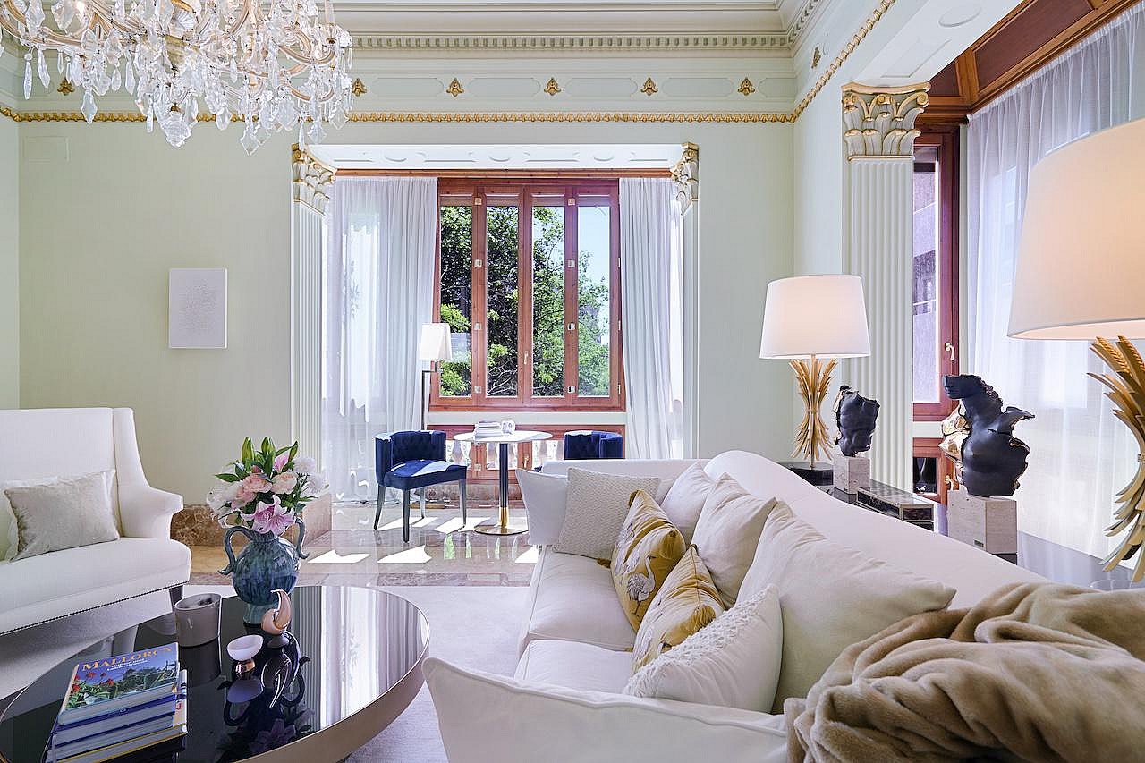 A formal living room with off-white sofas, a crystal chandelier, and ornate crown molding looking toward a windowed alcove.