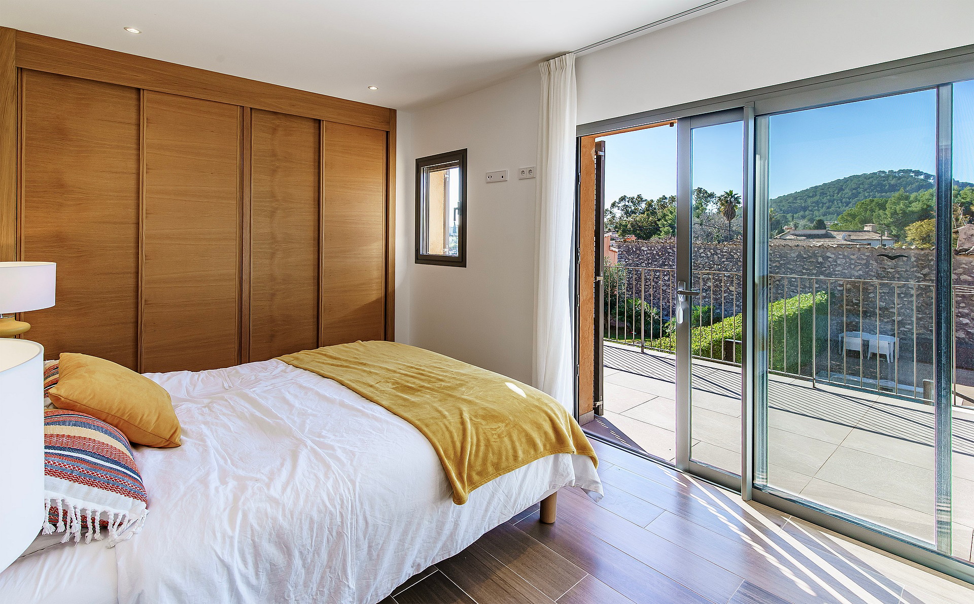 Bedroom interior with wooden floor-to-ceiling wardrobe, double bed, and wide sliding glass doors opening to a sunlit terrace.