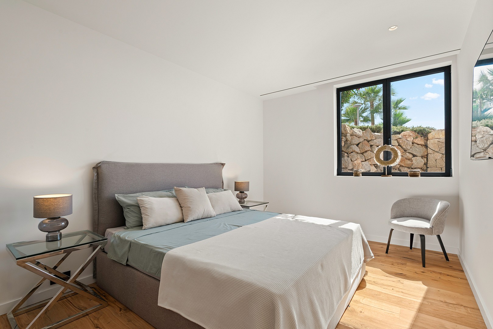 A minimalist bedroom featuring a grey upholstered bed, glass-topped side tables, and a view of a dry-stone wall outside.