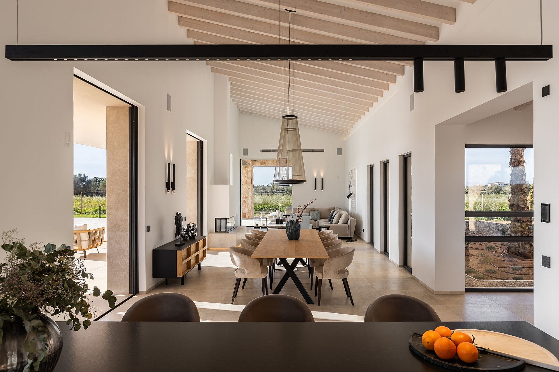 A spacious, bright open-plan room with white walls, wooden beamed ceilings, a large dining table, and direct terrace access.
