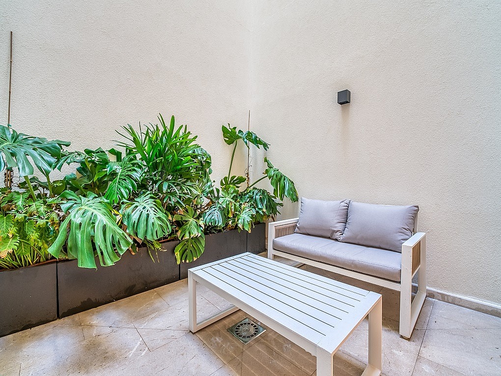 A small outdoor patio featuring a gray sofa, white coffee table, and large potted monstera plants against a beige wall.