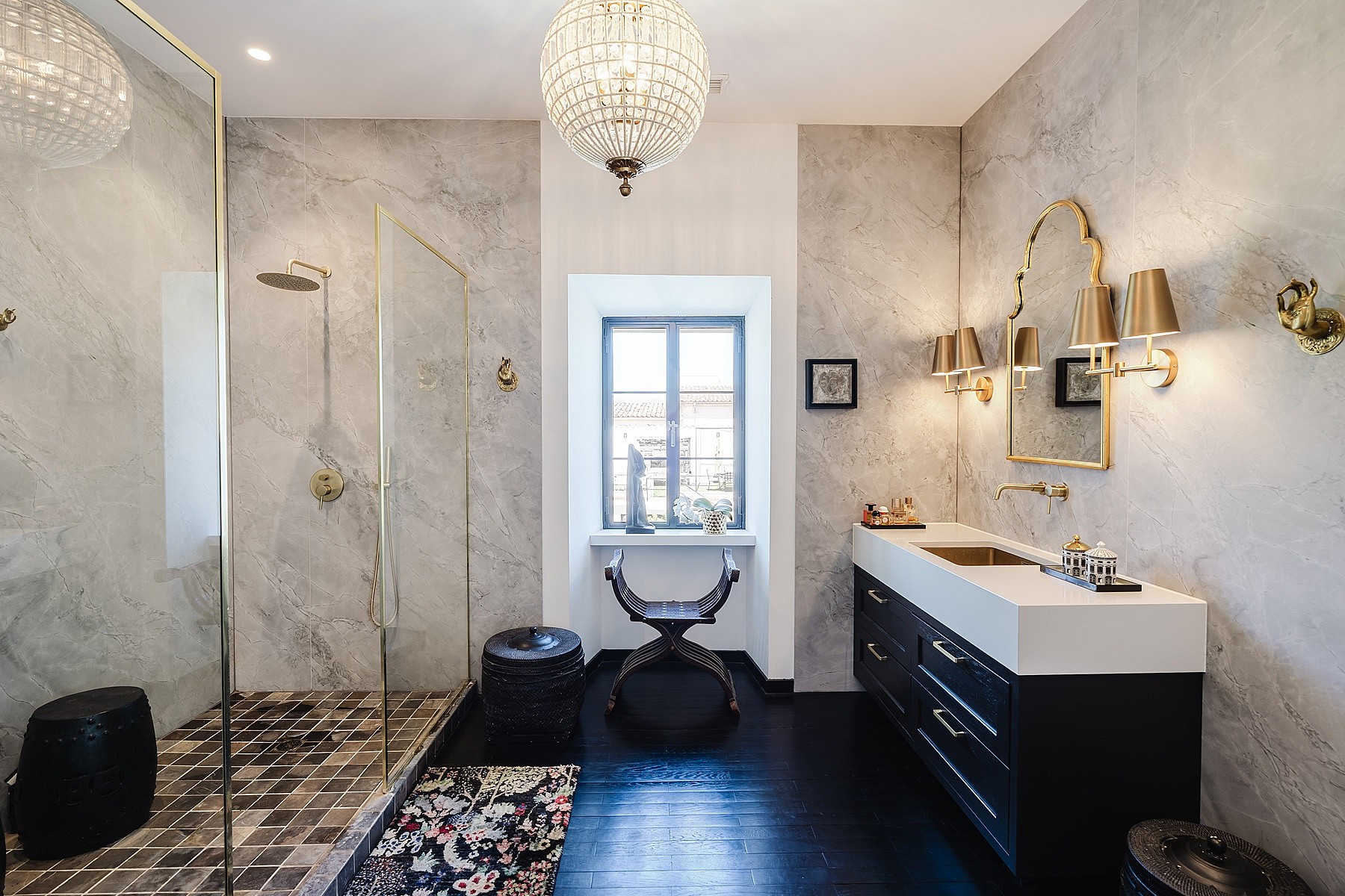 A modern bathroom featuring grey marble walls, a glass walk-in shower, a dark wood vanity with a stone basin, and gold accents.