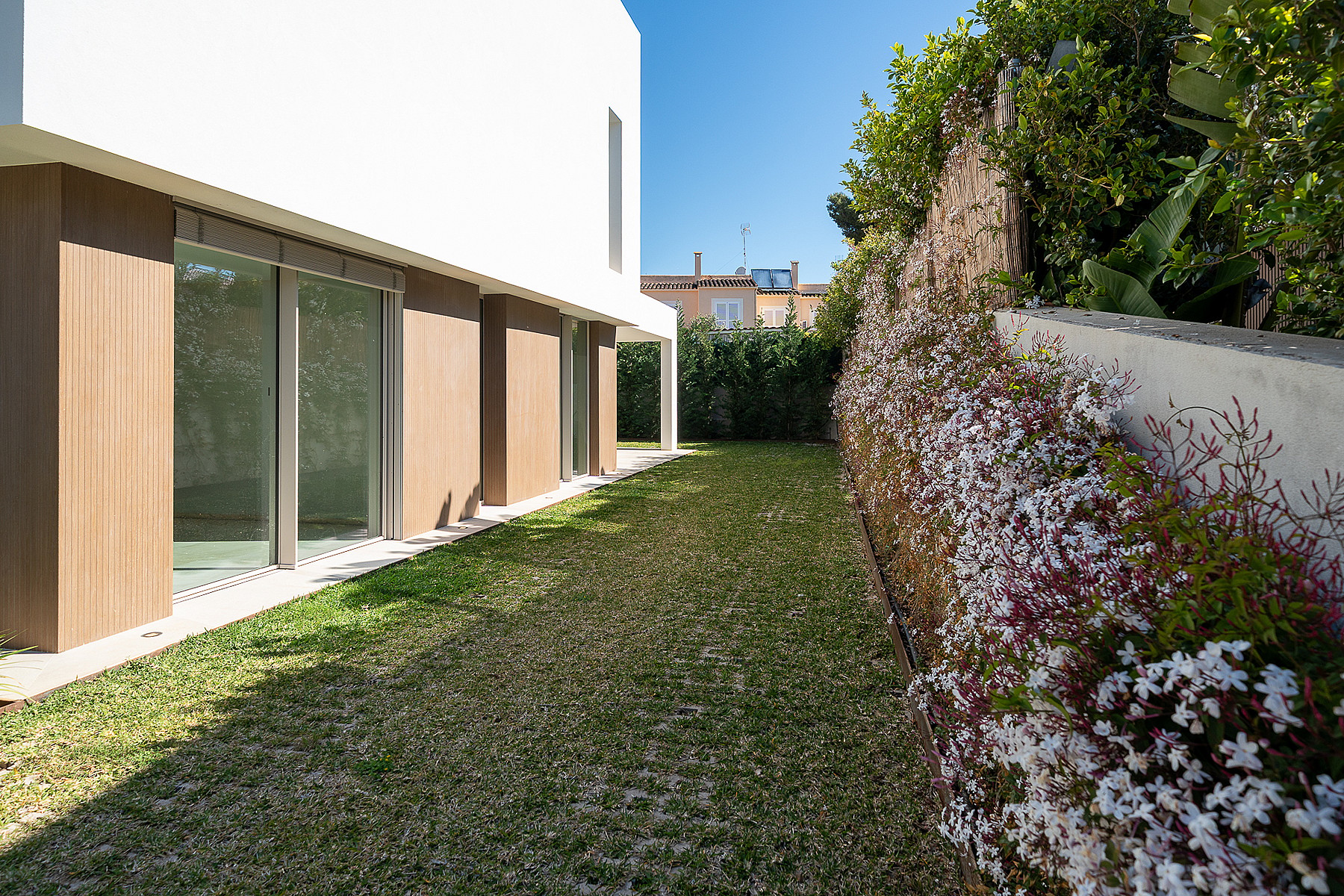 Side garden of a modern villa featuring a well-maintained lawn, white exterior wall with wood panels, and a blooming hedge.