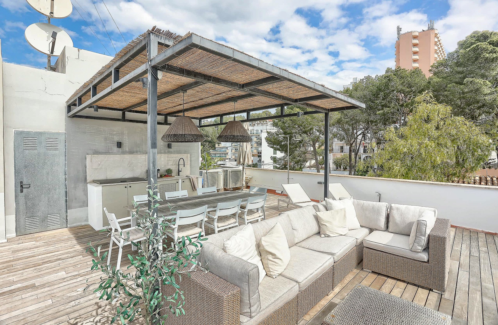 A rooftop terrace featuring a wicker sectional sofa, dining area, and outdoor kitchen under a wooden pergola.