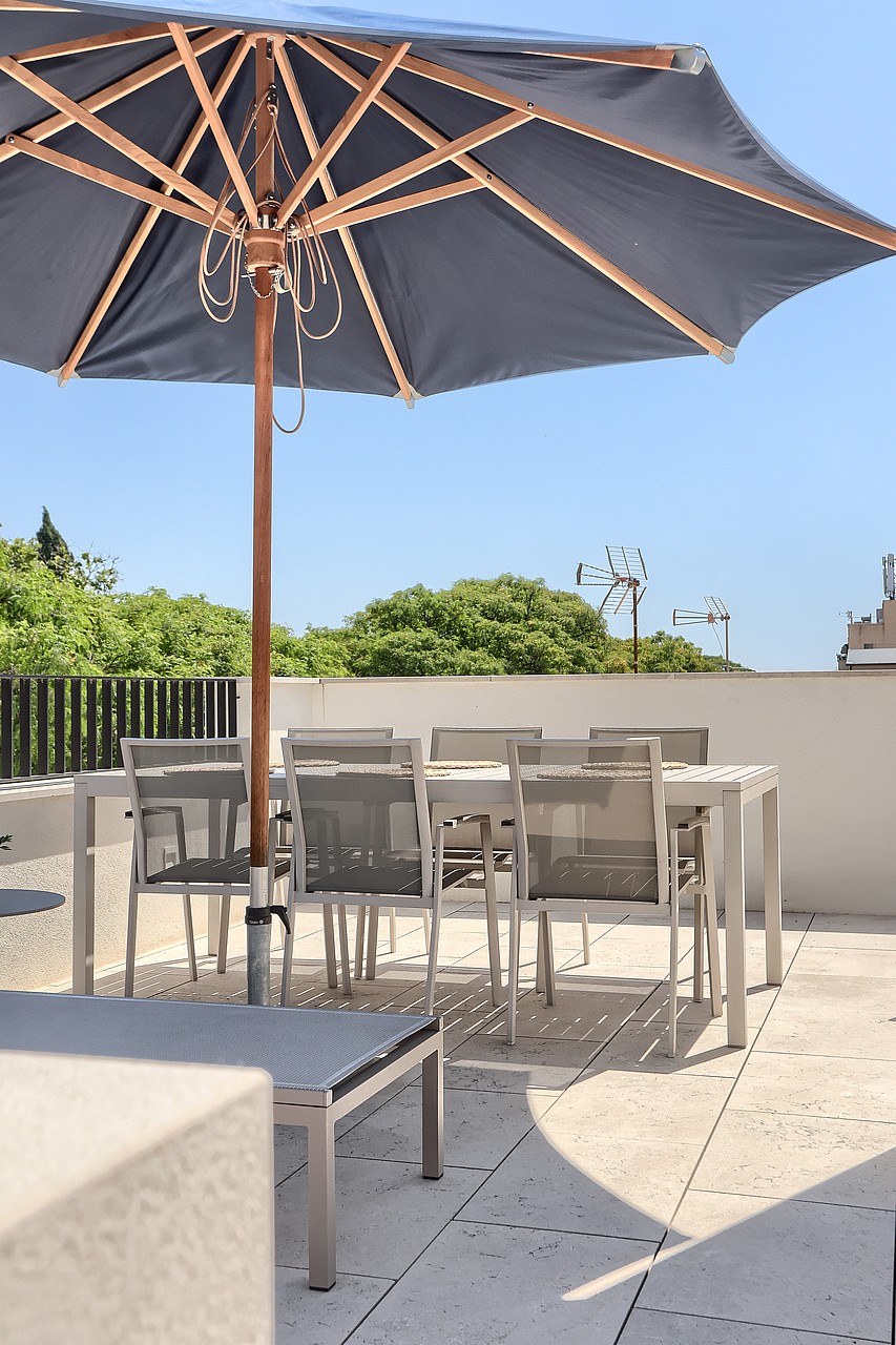 Rooftop terrace with a dining table and chairs shaded by a dark blue umbrella, set against a bright sky.
