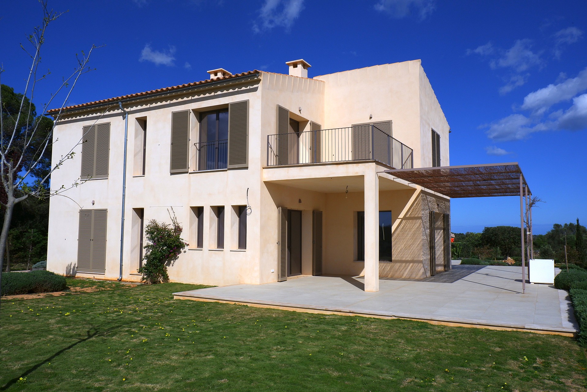 Exterior view of a cream-colored two-story finca with traditional wooden shutters, a shaded terrace, and a grassy garden.