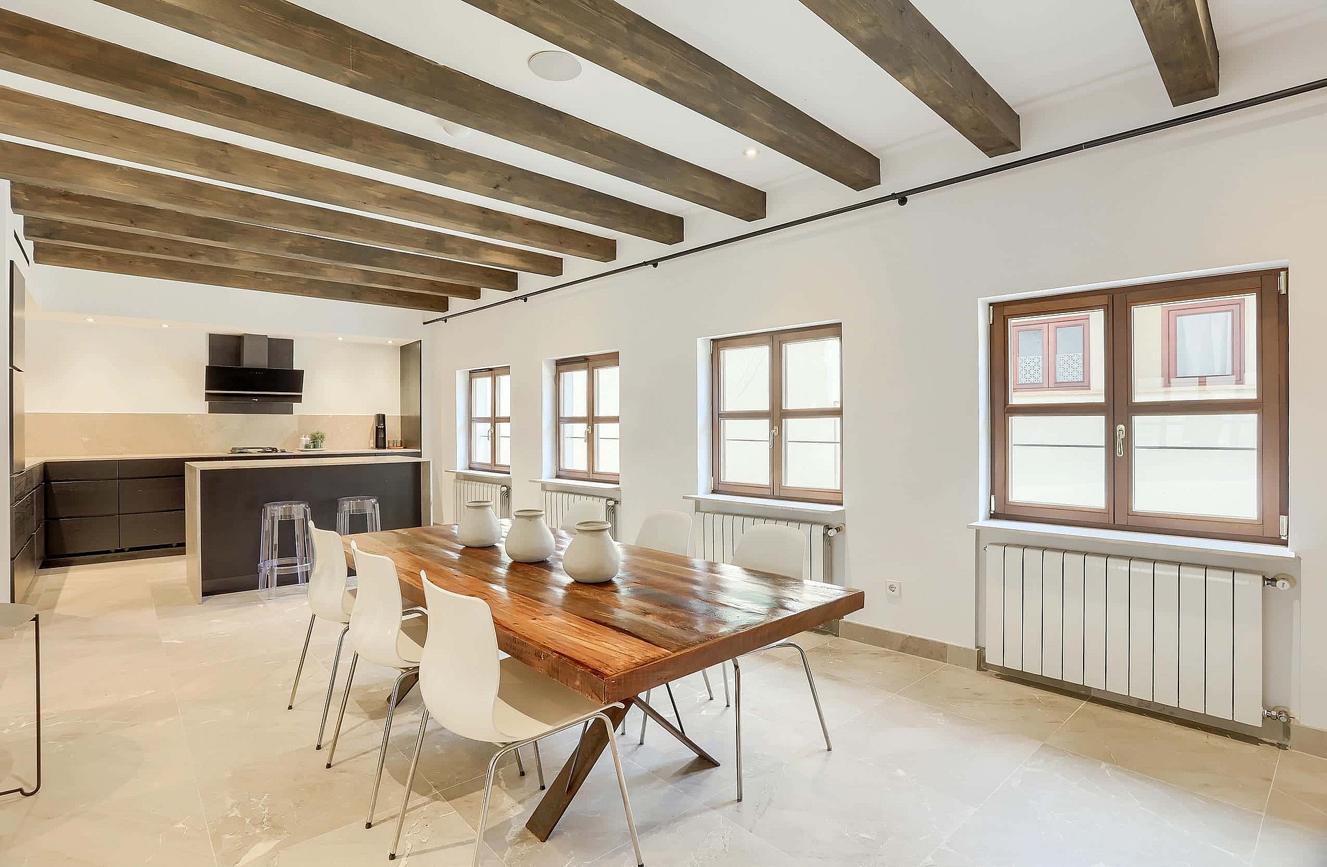 A bright open-plan kitchen and dining space with exposed wooden ceiling beams, a long wooden table, and minimalist white chairs.