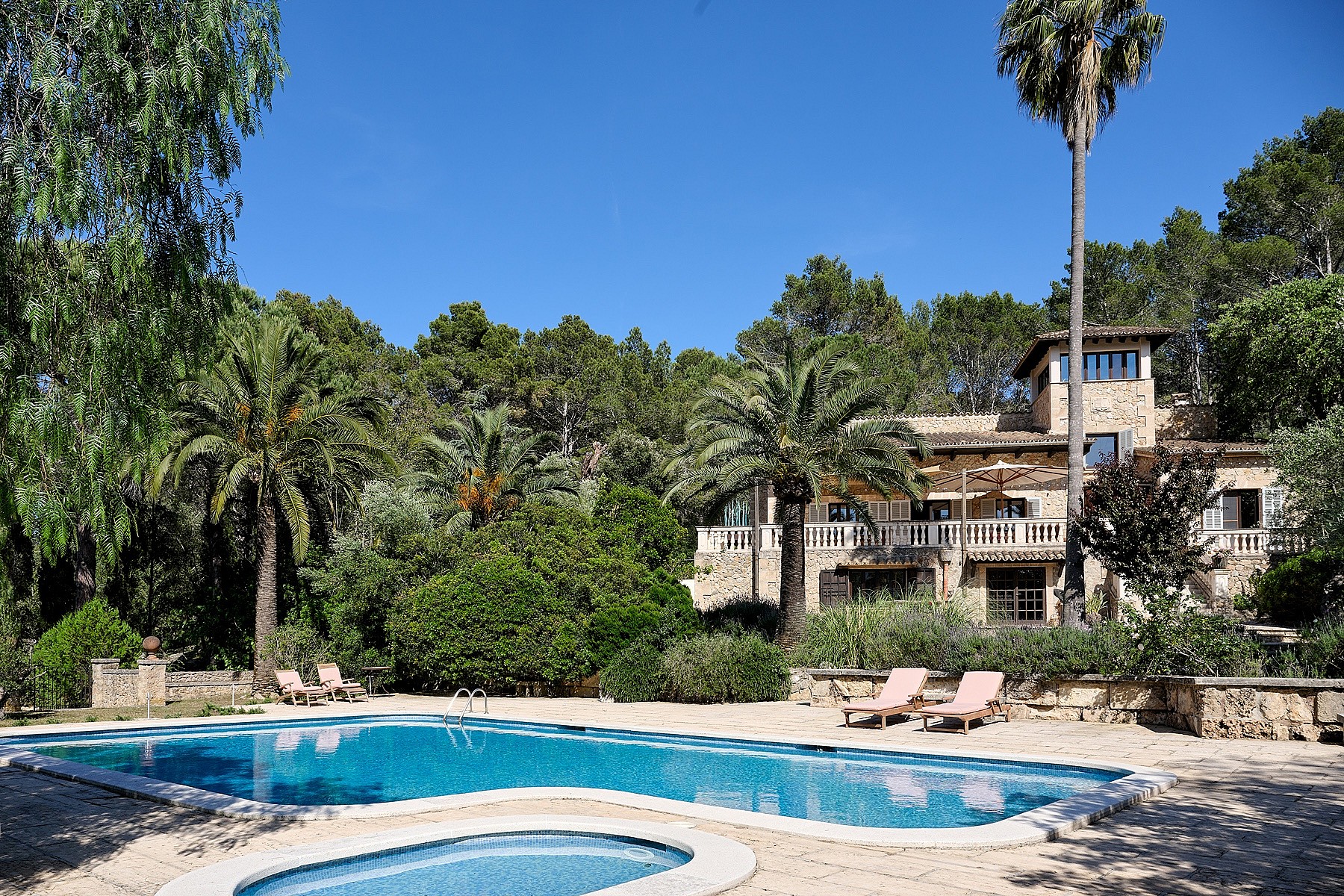 Exterior view of a stone finca with a large swimming pool, surrounding gardens, and mature palm trees under a clear sky.