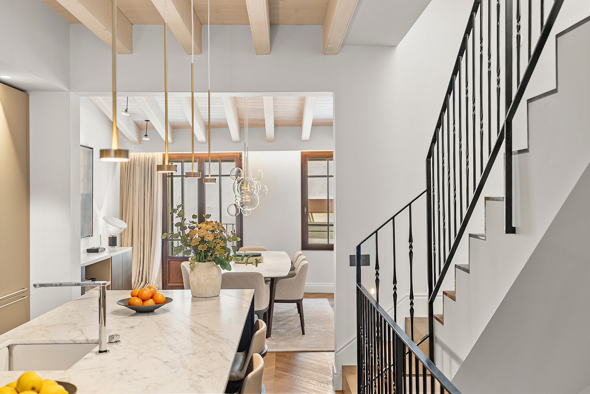 Interior view showing a black iron staircase railing, marble kitchen counter, and dining table under wooden ceiling beams.