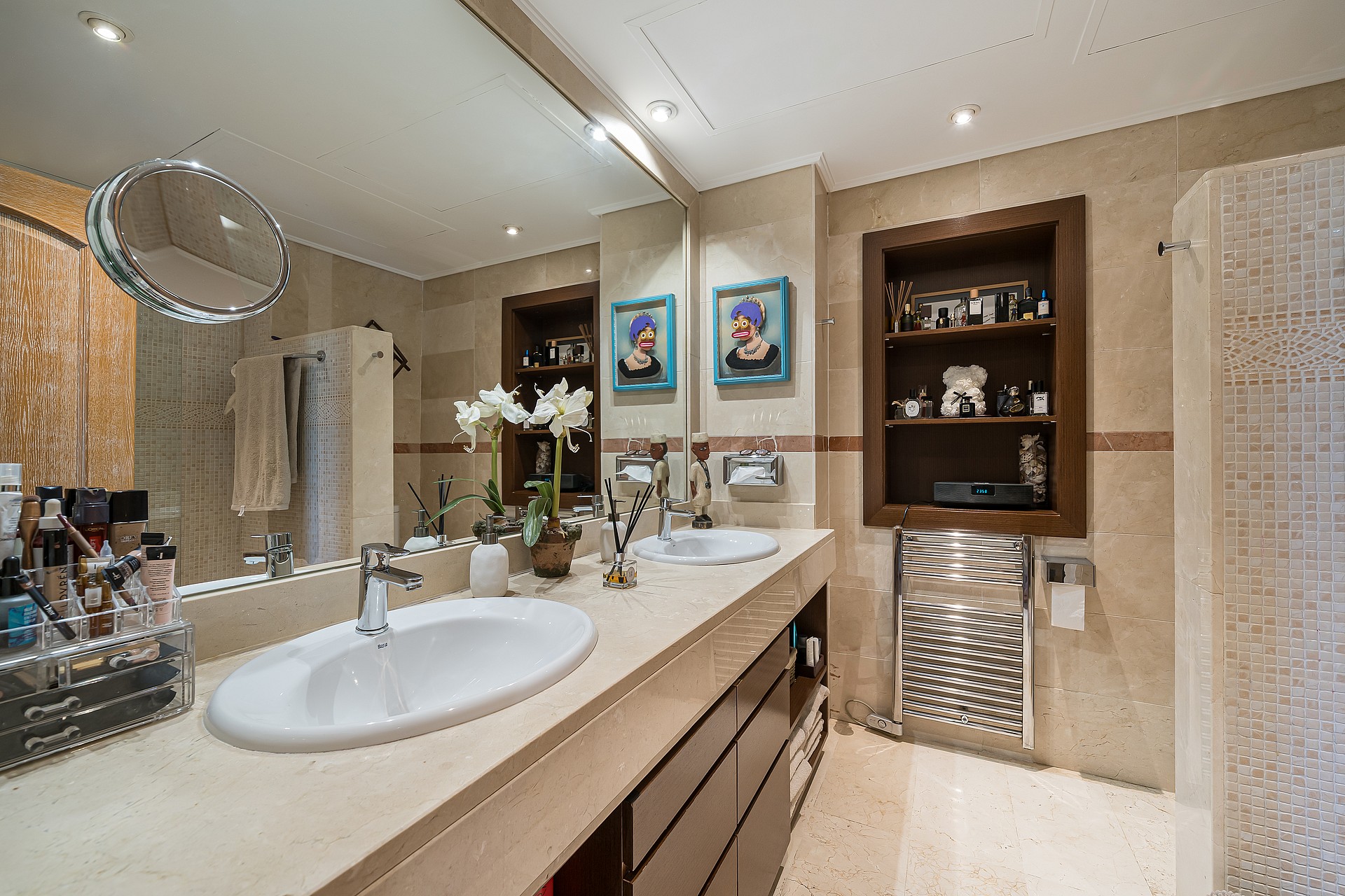 A master bathroom featuring a long stone-topped vanity with double sinks, a large wall-to-wall mirror, and recessed wall shelving.