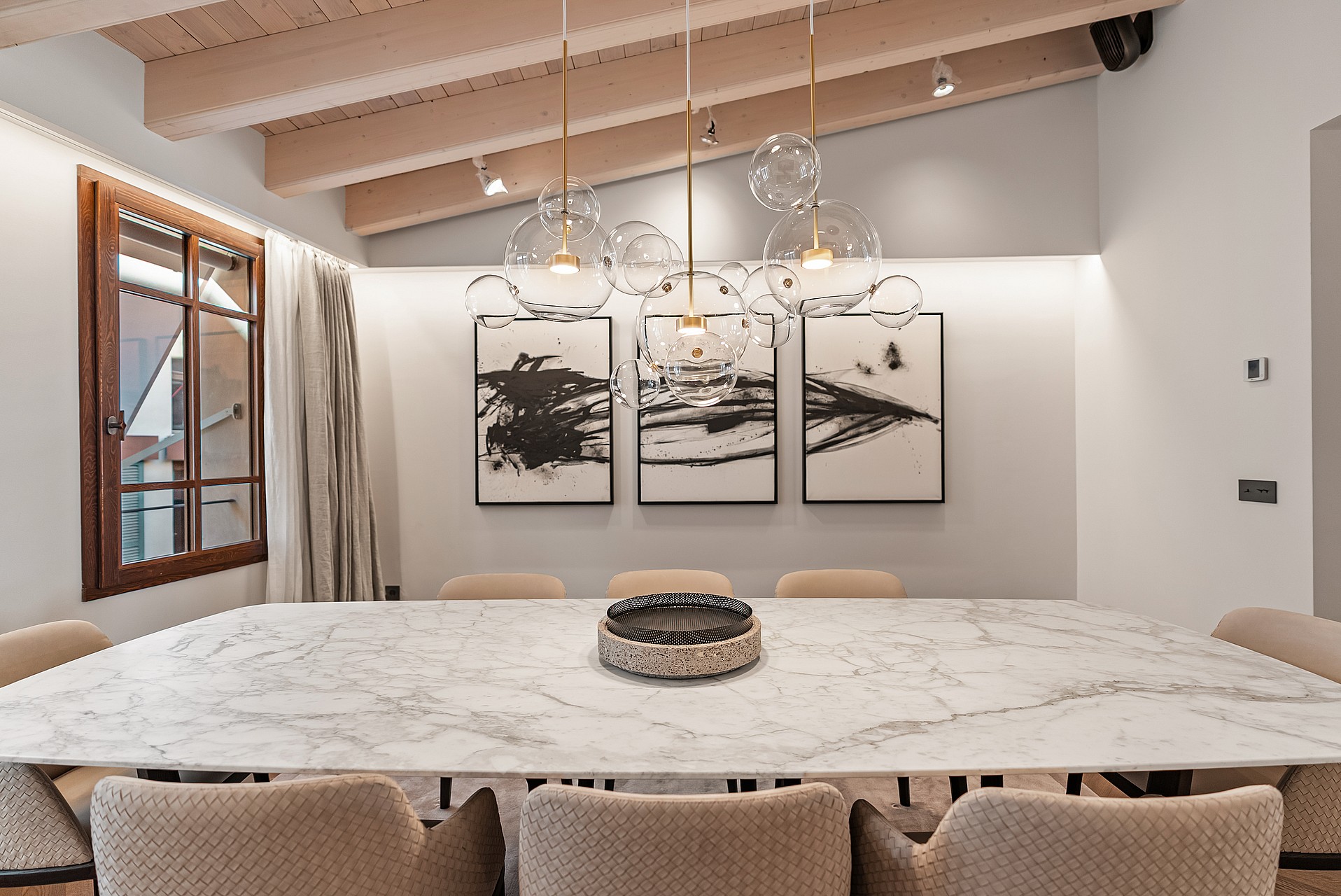 A modern dining space featuring a white marble table, glass globe pendant lights, and exposed light-wood ceiling beams.