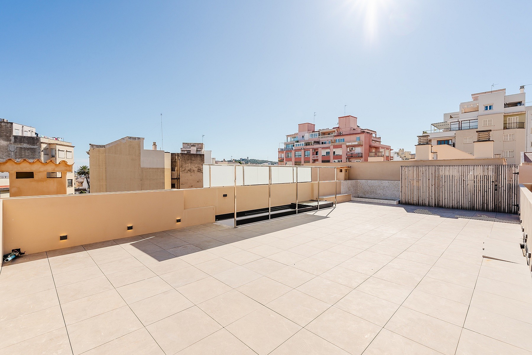 A wide, empty rooftop terrace with tiled flooring and low walls, overlooking surrounding city architecture under a clear blue sky.