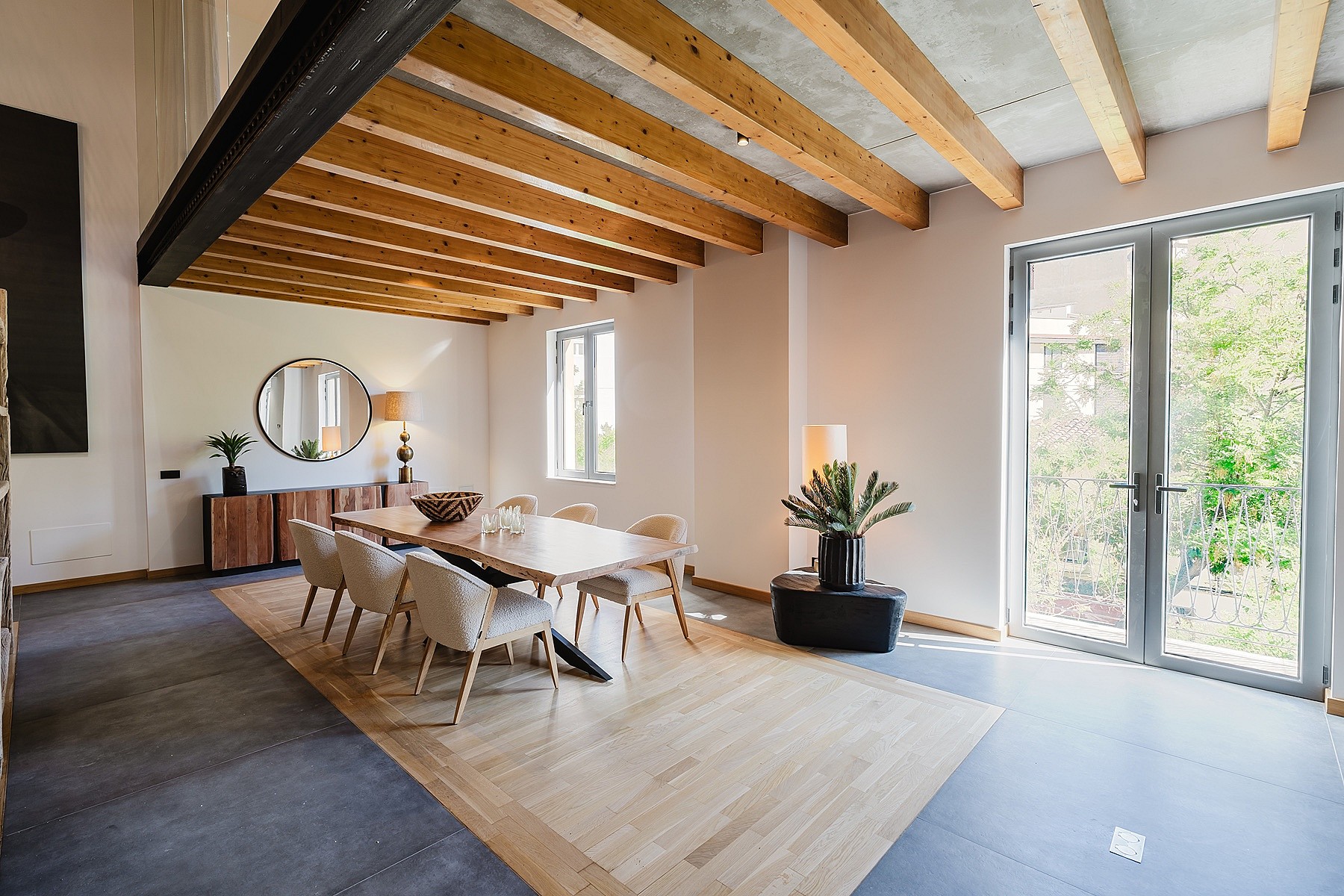 Dining area with wooden table, light-colored chairs, and exposed ceiling beams. Glass doors open to greenery.