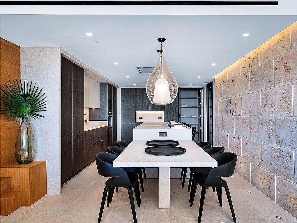 Modern open-plan kitchen with a large white island, dark cabinetry, and a light-colored natural stone feature wall.