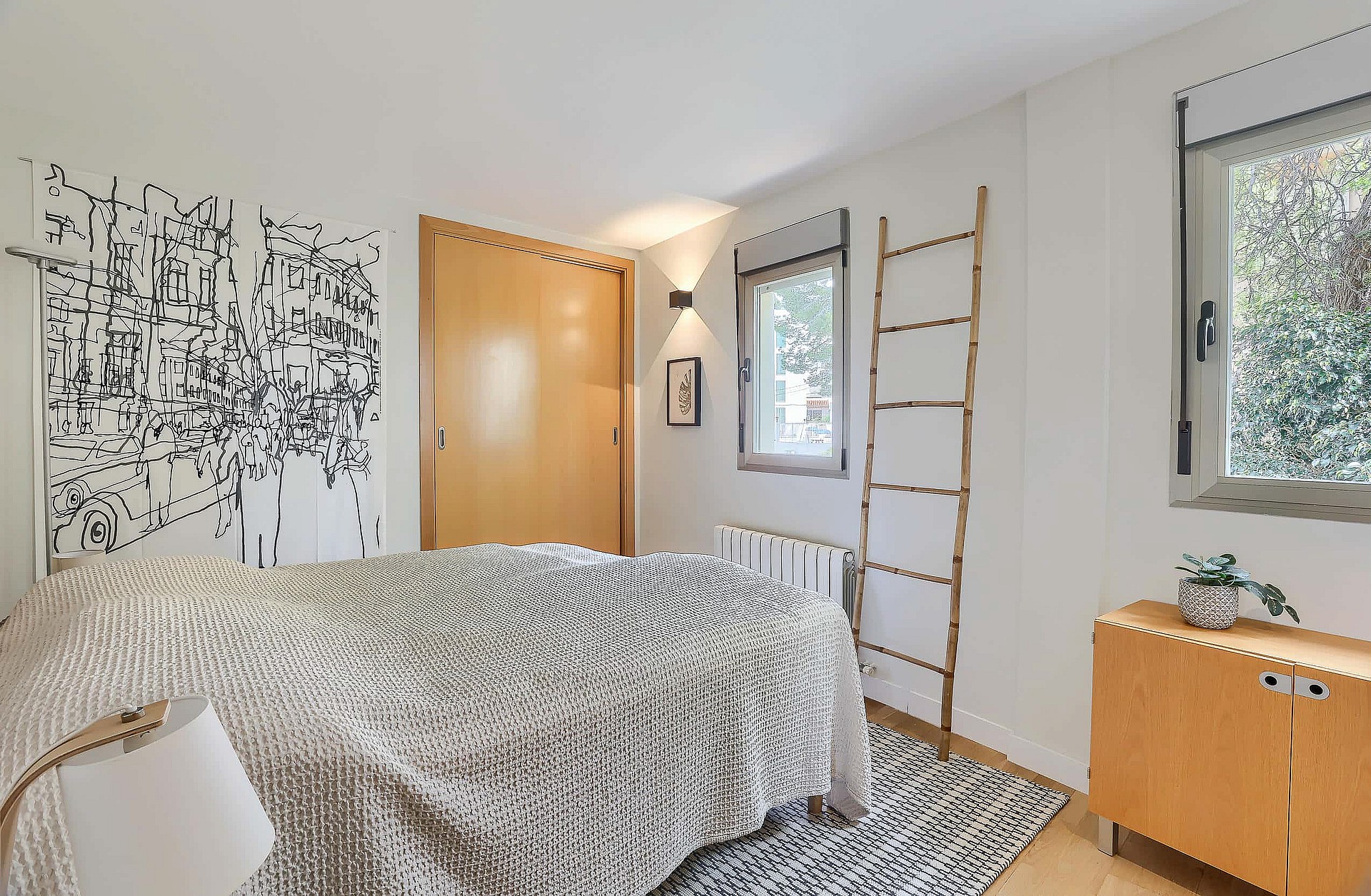 A minimalist bedroom featuring light wood closet doors, white walls, a patterned throw, and a decorative wooden ladder.