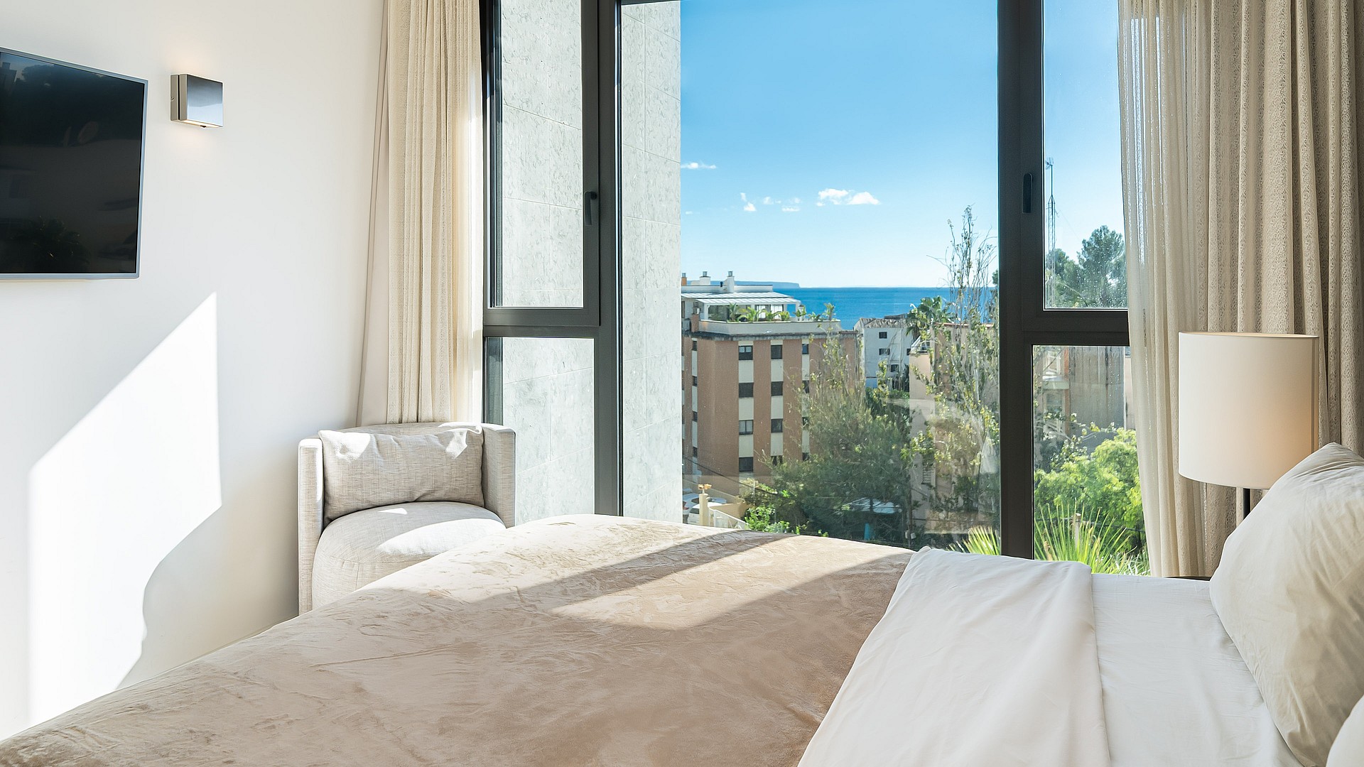 A partial view of a bedroom interior with a large window framing a sea view, featuring a tan bed cover and soft white curtains.