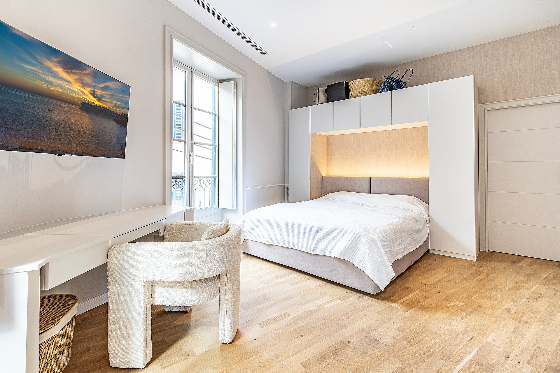 Modern bedroom with a white bed frame surrounded by built-in white cabinets, light oak wood flooring, and a textured white chair.