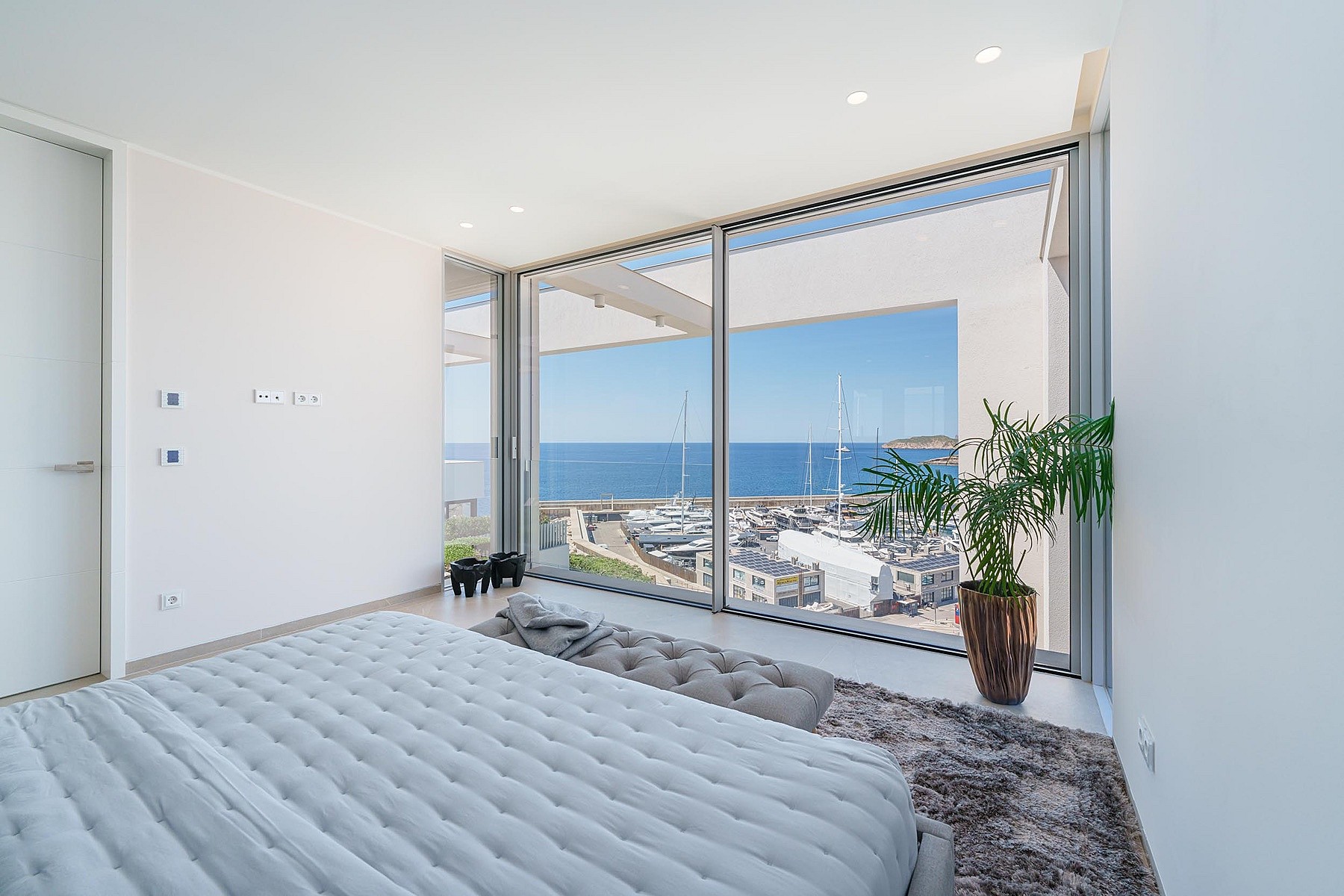 Contemporary bedroom with a quilted grey bed, full-height sliding glass walls, and a view of Port Adriano harbor.
