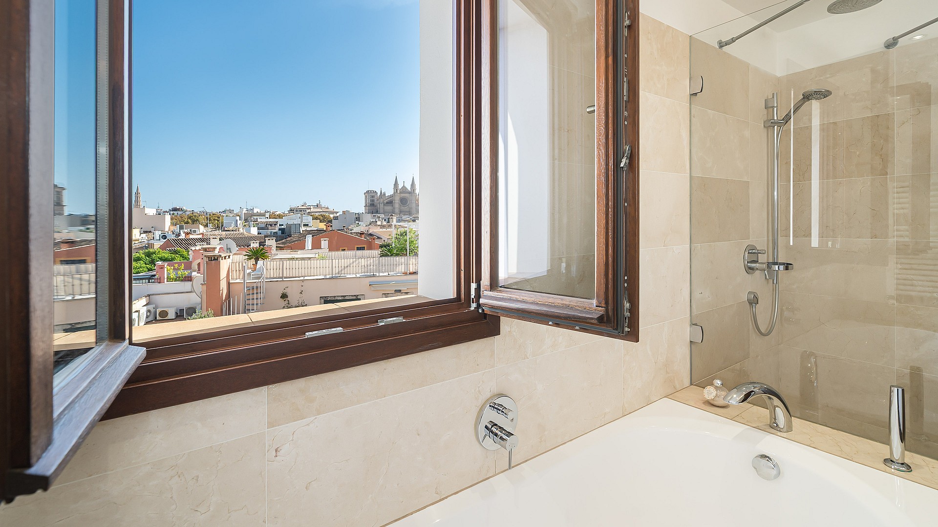 Modern bathroom interior featuring a white bathtub and glass shower next to an open window overlooking Palma's rooftops.