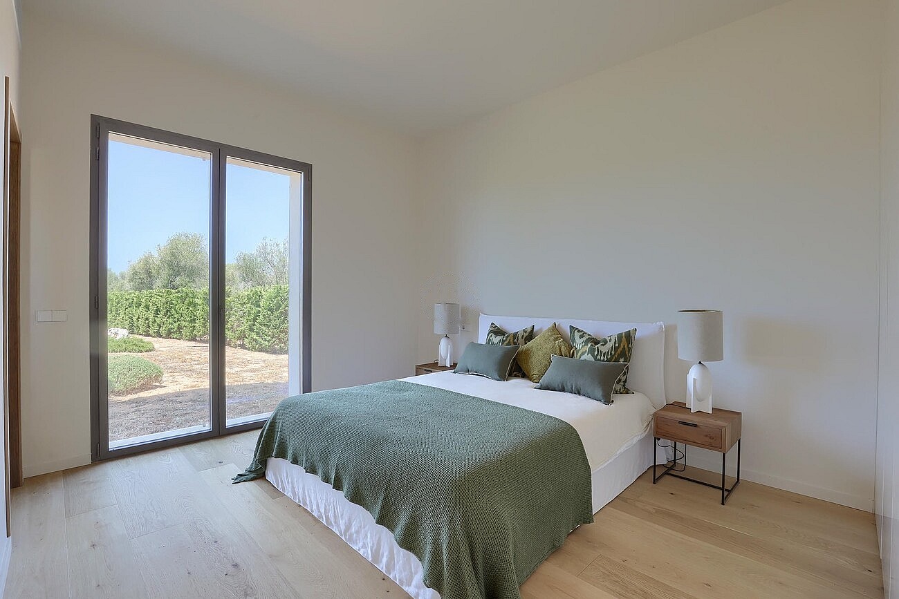 Bedroom with light wooden floors, a bed with a green spread, and a glass door opening to a terrace with garden views.