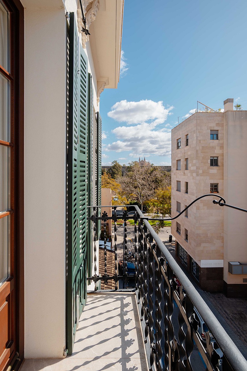 A narrow residential balcony featuring black decorative wrought iron railings and green shuttered doors overlooking a street.