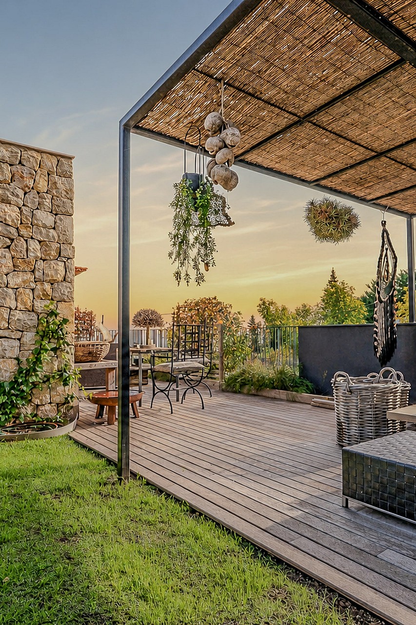 Covered outdoor terrace with wooden decking, stone wall, and metal pergola framing mountain views at sunset.