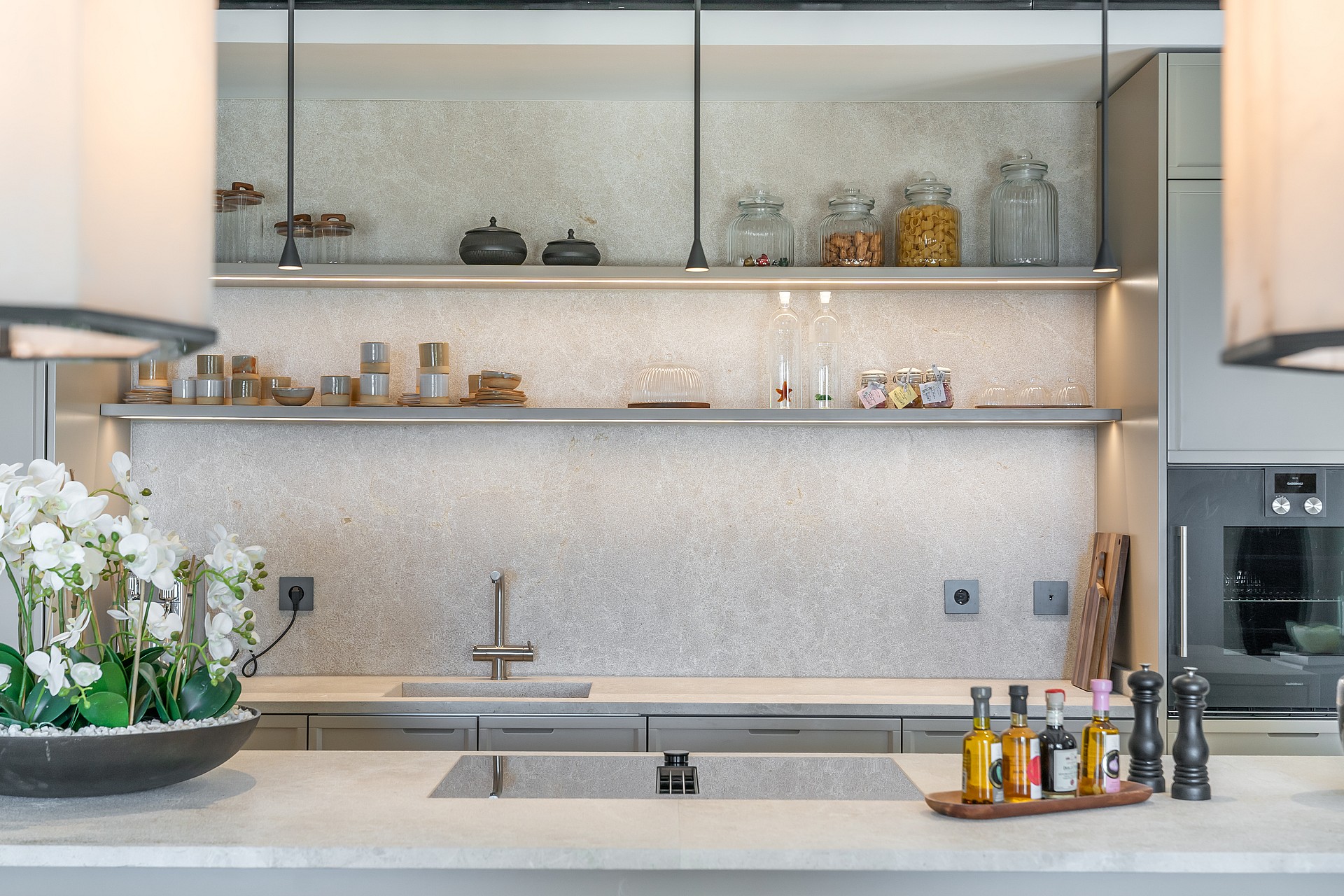 A contemporary kitchen showing a textured stone backsplash, slim floating shelves with LED lighting, and grey cabinetry.