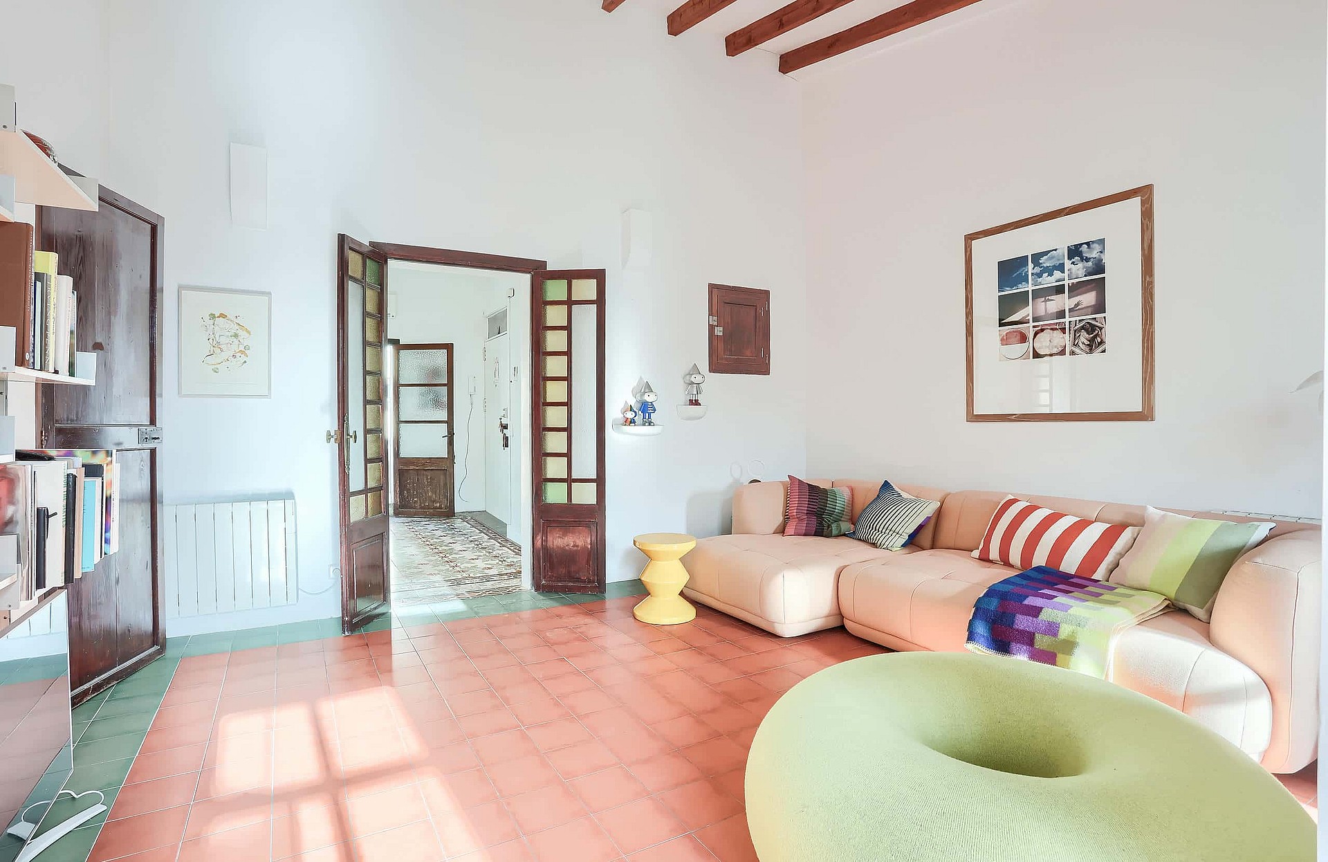 A bright living room with reddish terracotta tile floors, white walls, and a large sectional sofa. Wooden exposed ceiling beams are visible above. A set of open glass-paneled doors leads to an adjacent room.