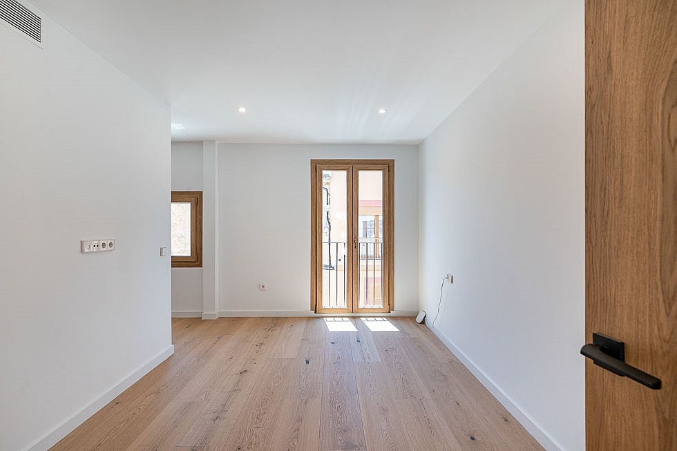 A bright, empty bedroom with light oak wood floors, white walls, and a wooden framed glass door leading to a small balcony.