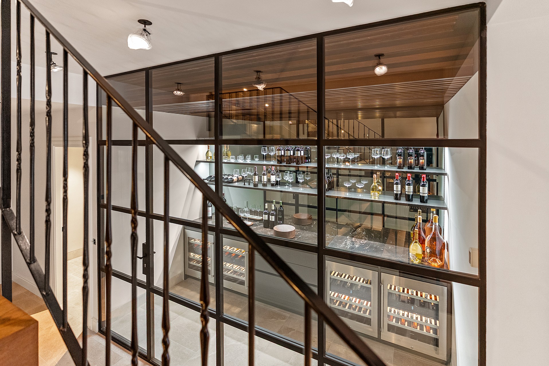 Wrought iron staircase railing in front of a climate-controlled glass wine cellar with internal shelving and lighting.