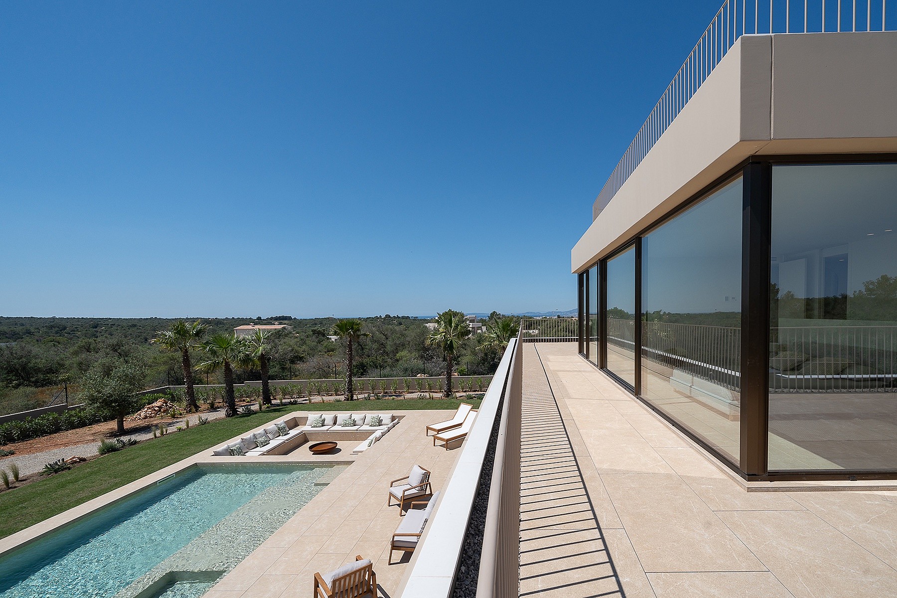 An elevated terrace with glass railings overlooking a swimming pool, sun loungers, and distant mountain landscape.