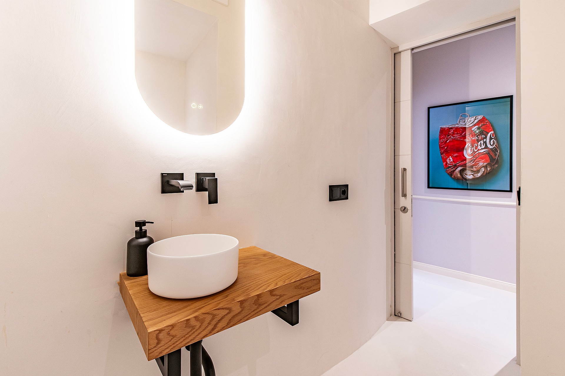 A modern guest bathroom featuring a wall-mounted wooden vanity, a white vessel sink, black wall-mounted faucet, and backlit mirror.