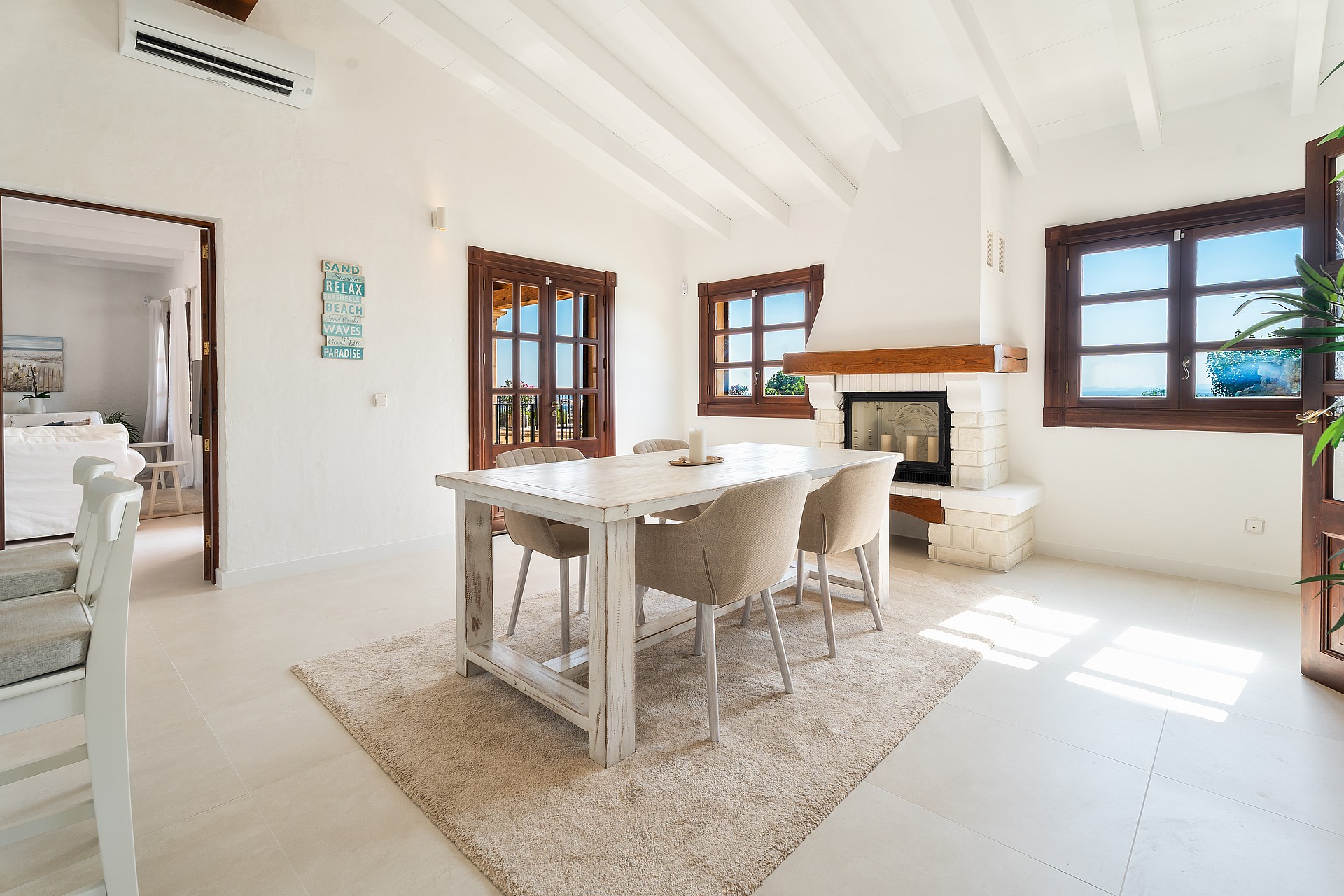 Zona de comedor con mesa de madera, sillas de mimbre y chimenea de piedra sobre muros blancos.
