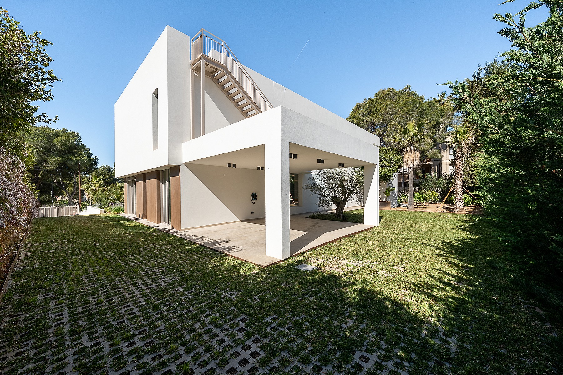 Side view of a white, two-story modern villa with a covered terrace, external metal staircase, and a green grass garden.
