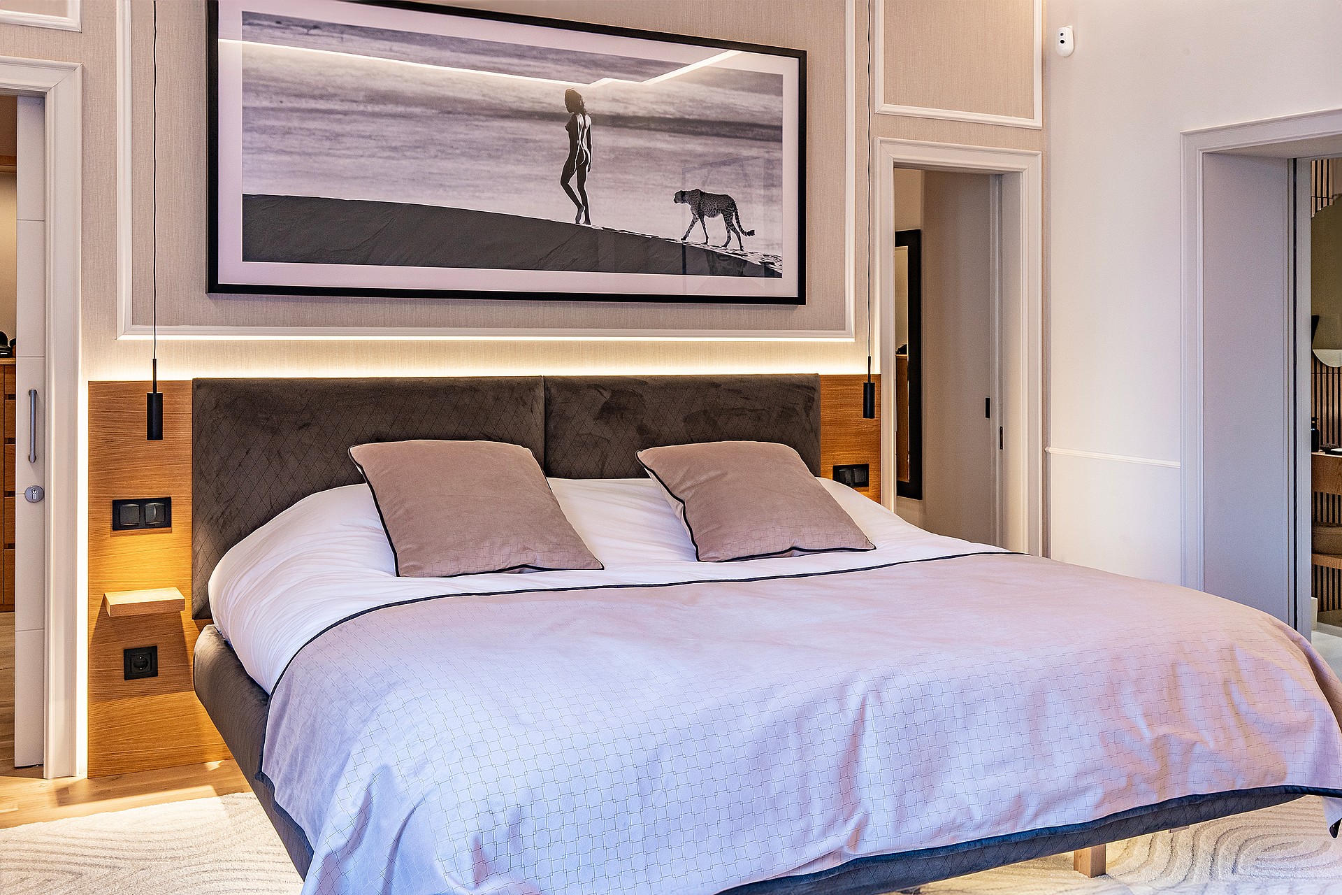 A modern master bedroom featuring a grey upholstered headboard, wooden paneling, and an oversized framed art piece above.