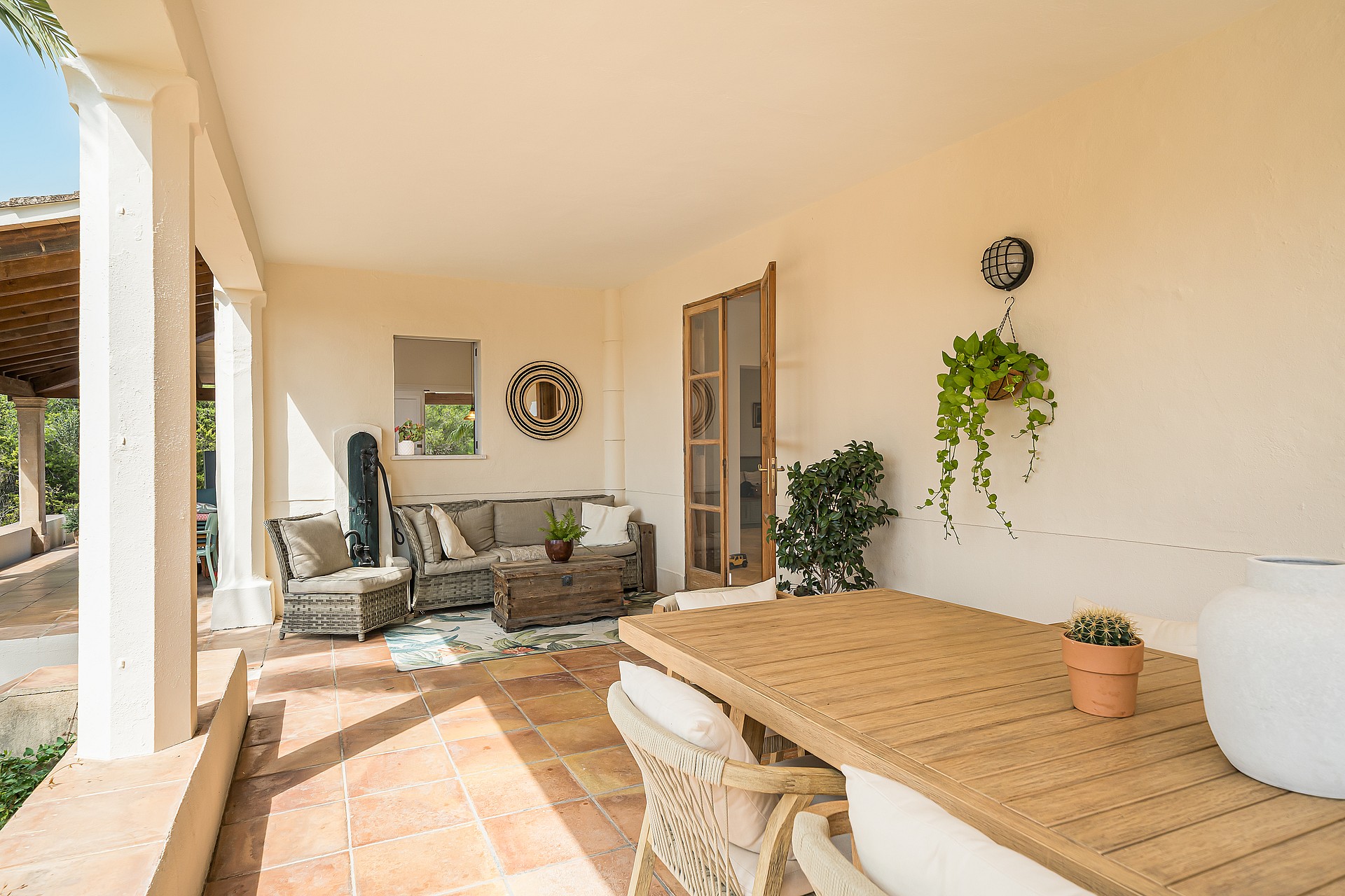 Covered outdoor terrace with terracotta tile flooring, wicker lounge seating, and a solid wood dining table in sunlight.