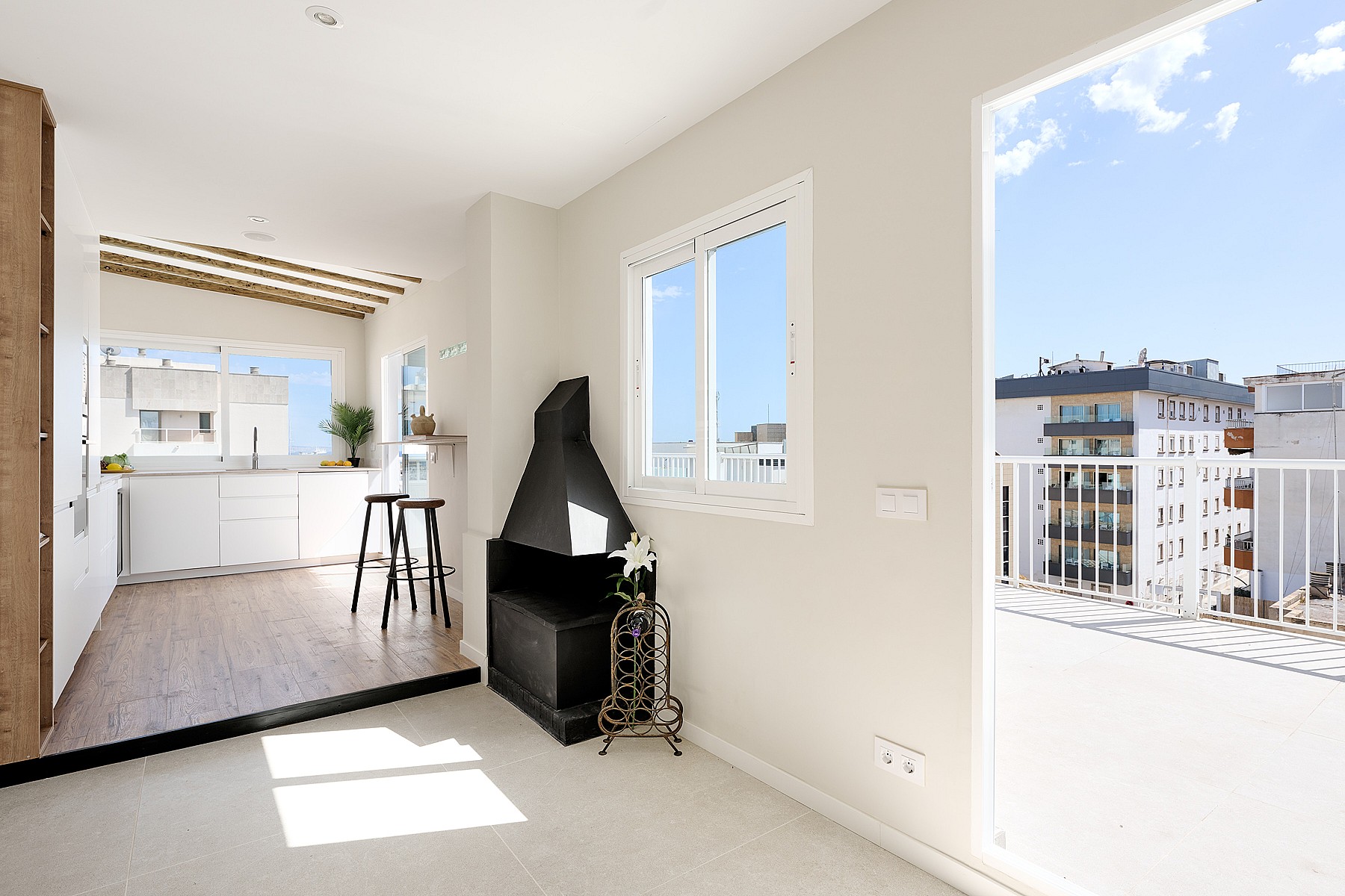 Bright open-plan living space featuring a black conical fireplace, white walls, and a glass door opening to a sunlit balcony.