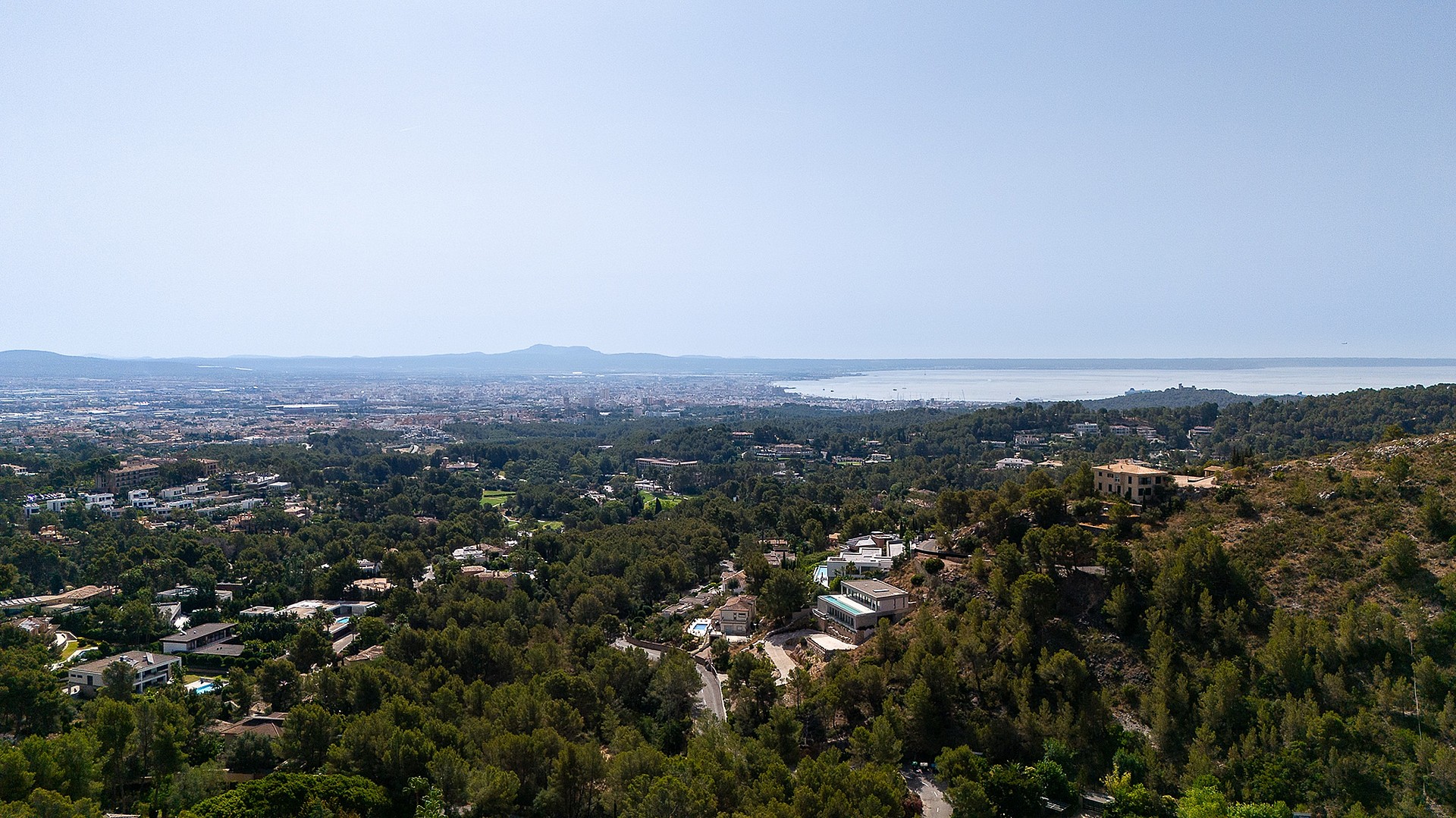High-angle aerial view of the Son Vida landscape showing expansive wooded hills sloping towards the distant coastline and sea.