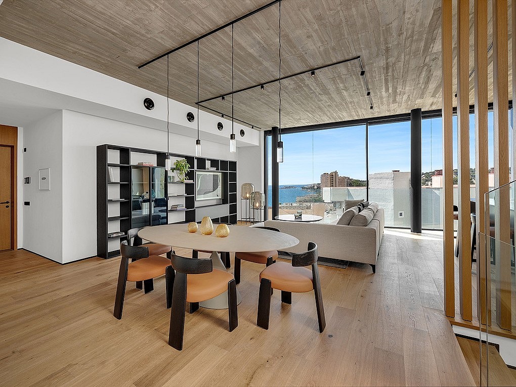 Modern open-plan living and dining room with concrete ceiling, wooden floors, and floor-to-ceiling windows overlooking the sea.