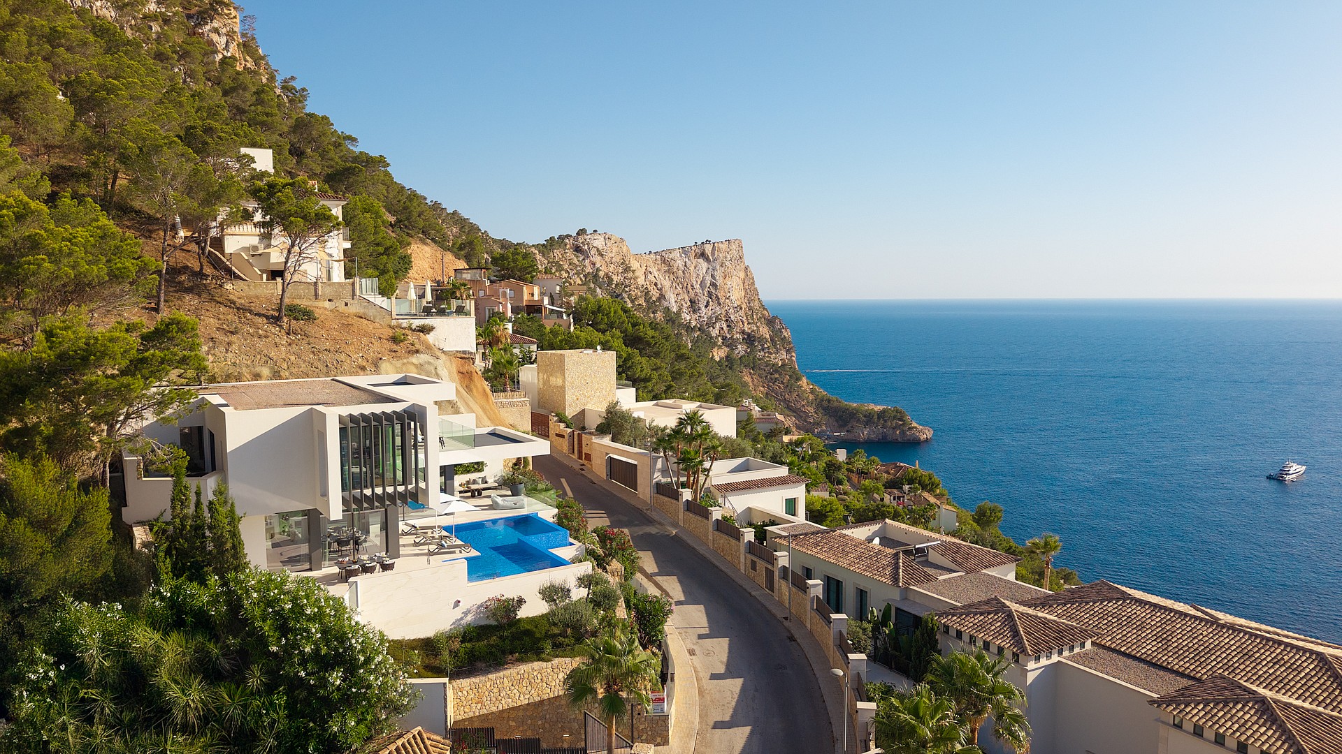 Aerial view of a white cliffside villa with a blue swimming pool, situated along a coastal road overlooking the Mediterranean.