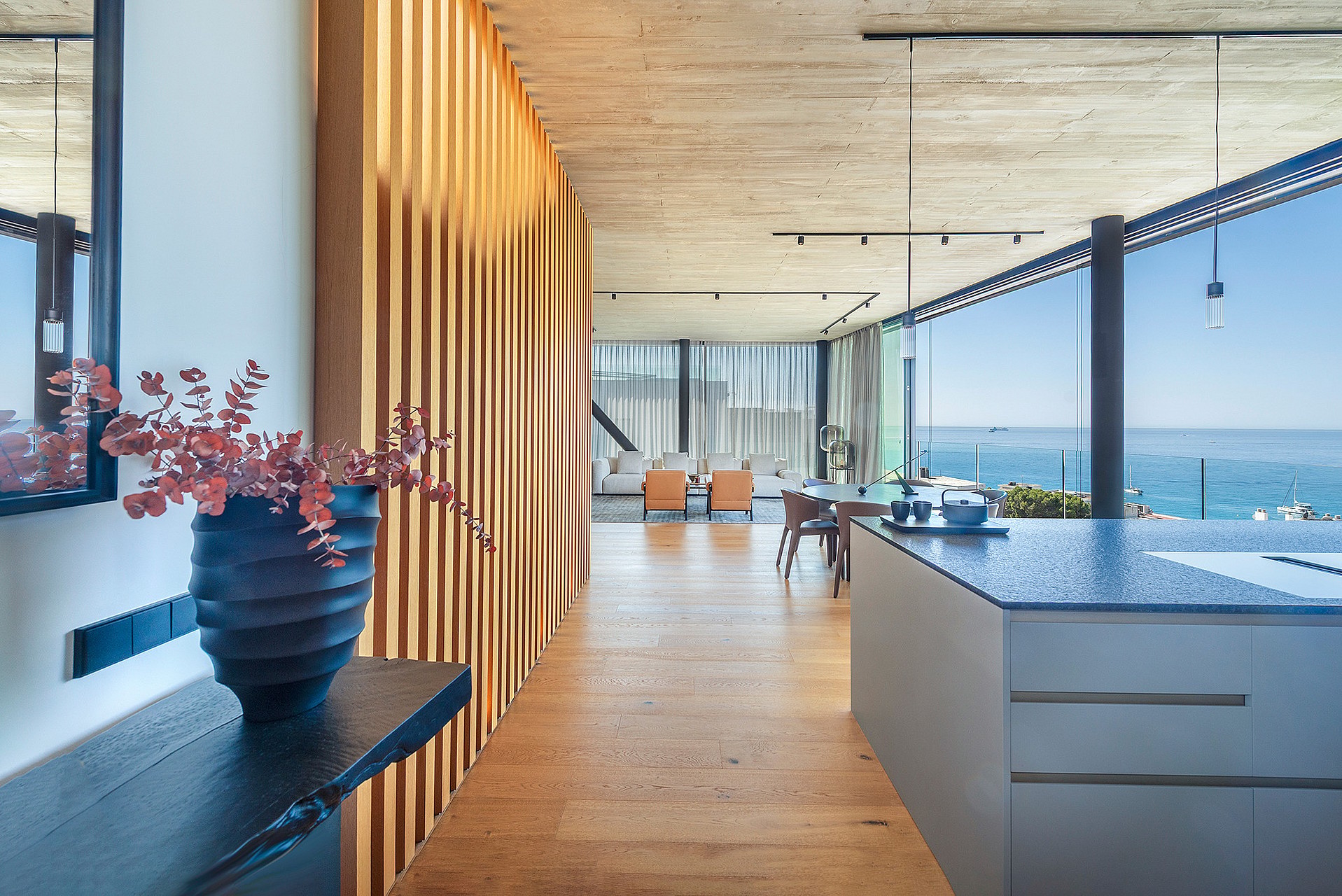 Interior of a modern kitchen showing a textured wood slat wall divider, dark kitchen island, and open-plan living area with horizon sea views.